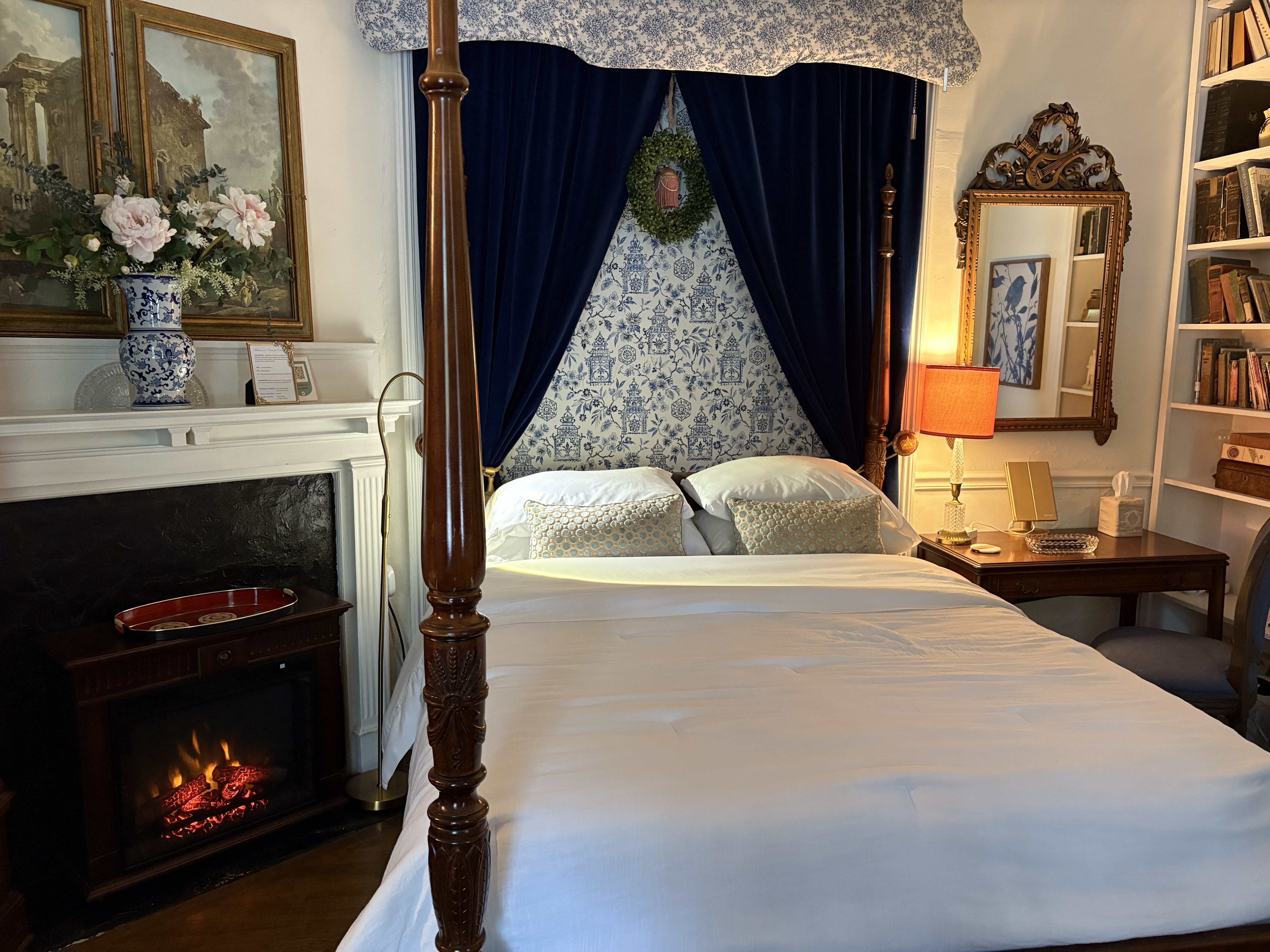 A traditional, cozy bedroom featuring a four-poster wooden bed with white linens, set against a backdrop of ornate blue and white patterned wallpaper and deep blue velvet curtains hanging from a matching valance. A tall white bookshelf filled with books, an ornate gold mirror, and a small desk with an orange lamp sit to the right of the bed.