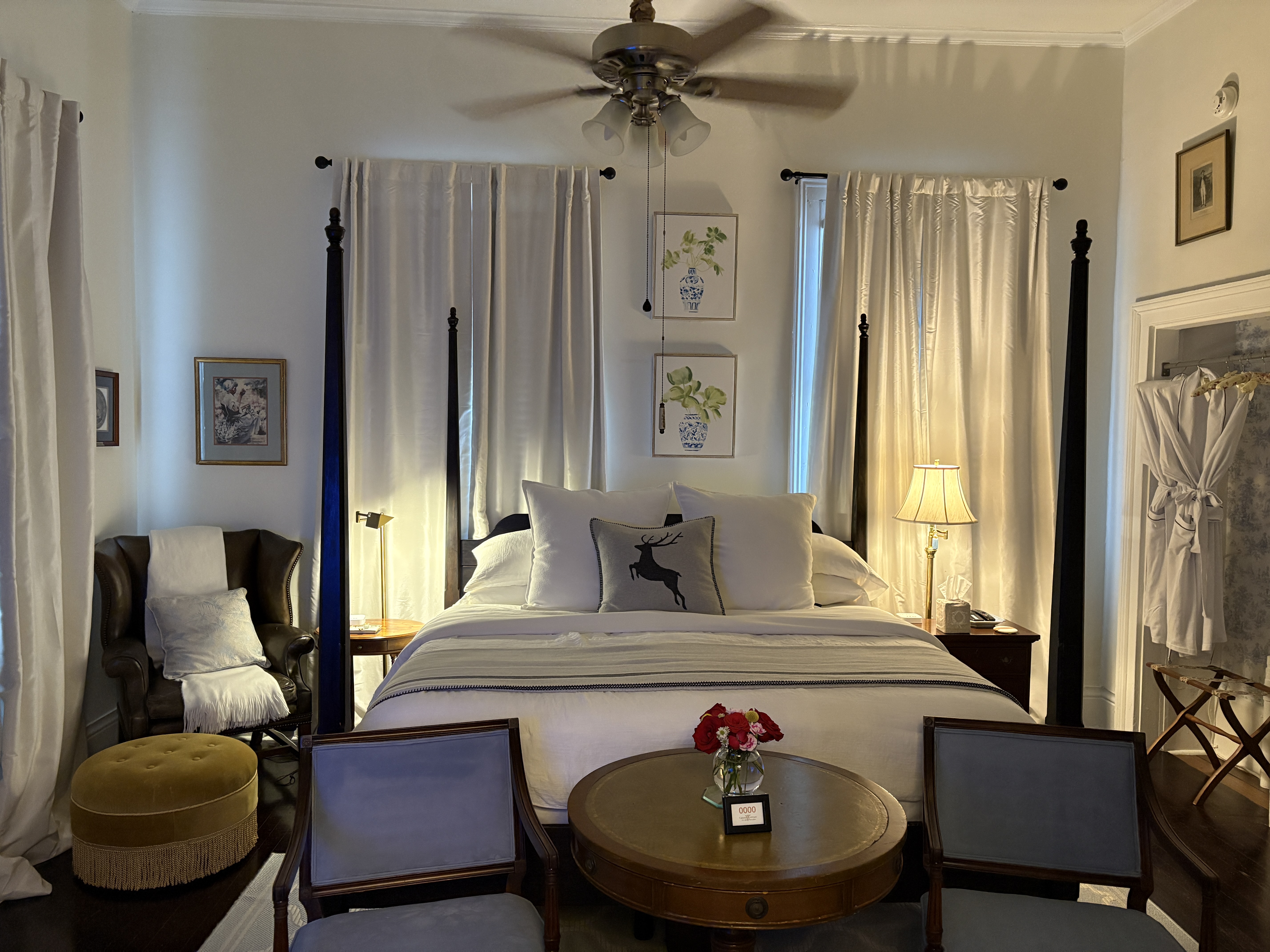 A symmetrical shot of a bedroom featuring a dark four-poster bed with white linens and a grey throw. In the foreground, two small blue chairs flank a round wooden table with a vase of red roses, while a black leather wingback chair with a yellow ottoman sits to the left. The room is decorated with framed botanical prints and two windows draped in long white curtains.