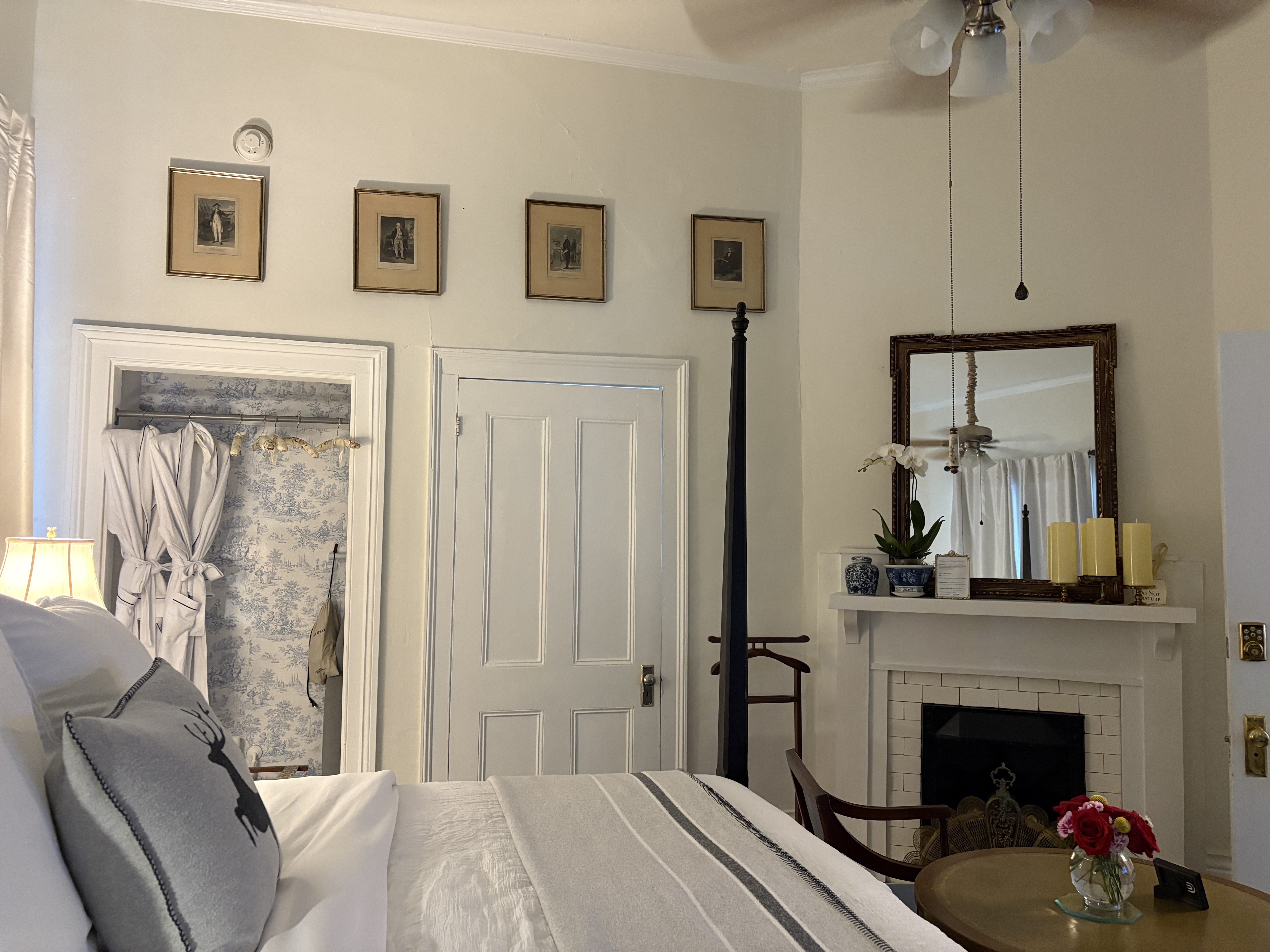 An elegant bedroom featuring a dark wood four-poster bed with crisp white linens and a grey throw blanket. In the foreground, two light blue chairs face a round wooden table topped with a small vase of red flowers, while a dark leather wingback chair with a yellow ottoman sits to the left. The room is accented with long white curtains framing the windows and several framed botanical prints on the walls.