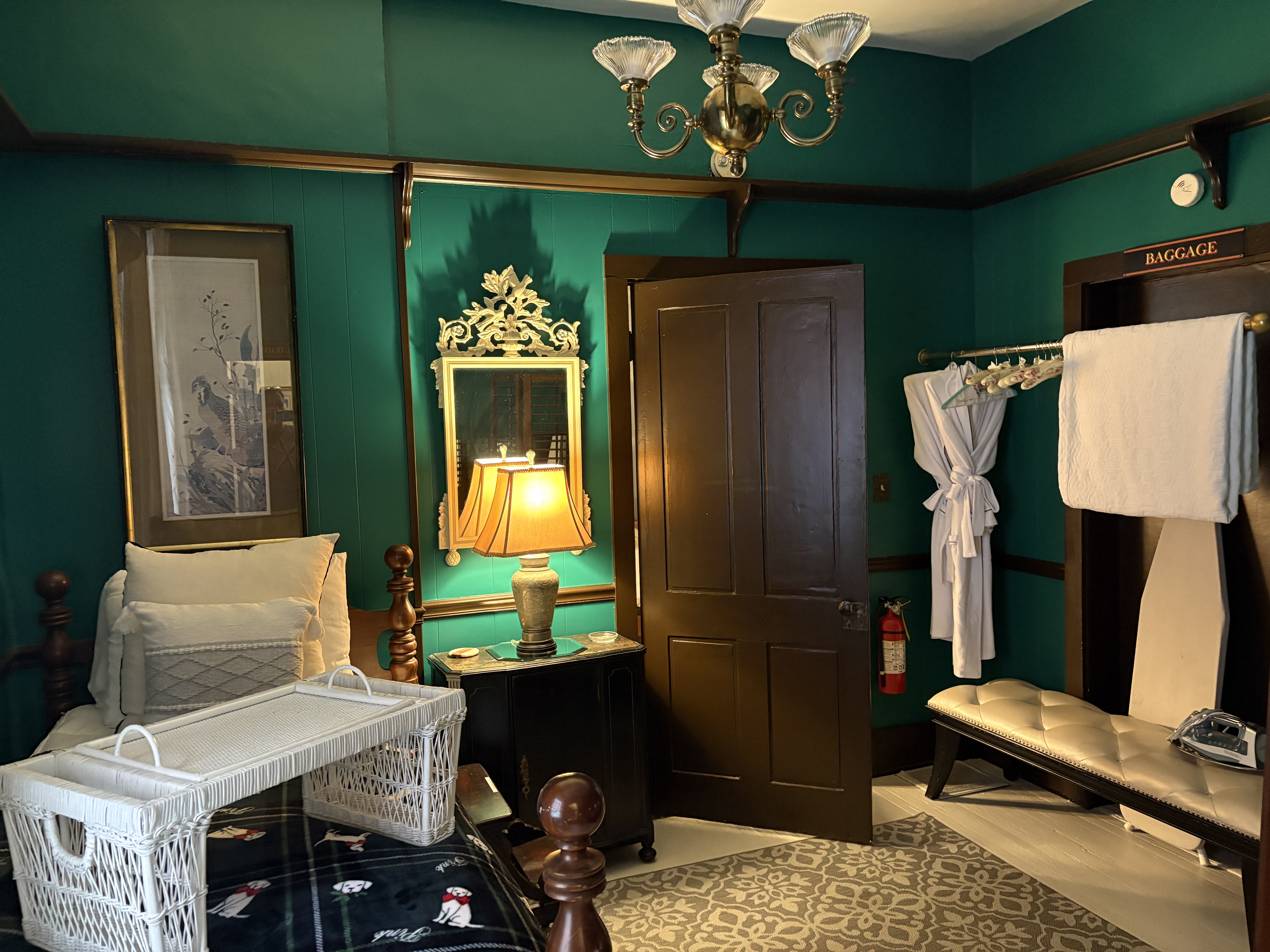 A bedroom with deep teal walls and dark wood accents features a bed with white pillows and a white wicker breakfast tray. To the right of a closed wooden door, a white bathrobe and towels hang on a rack next to a padded bench and an ironing board. An ornate gold mirror and a lamp with a warm glow are positioned on a small black nightstand to the left of the door.