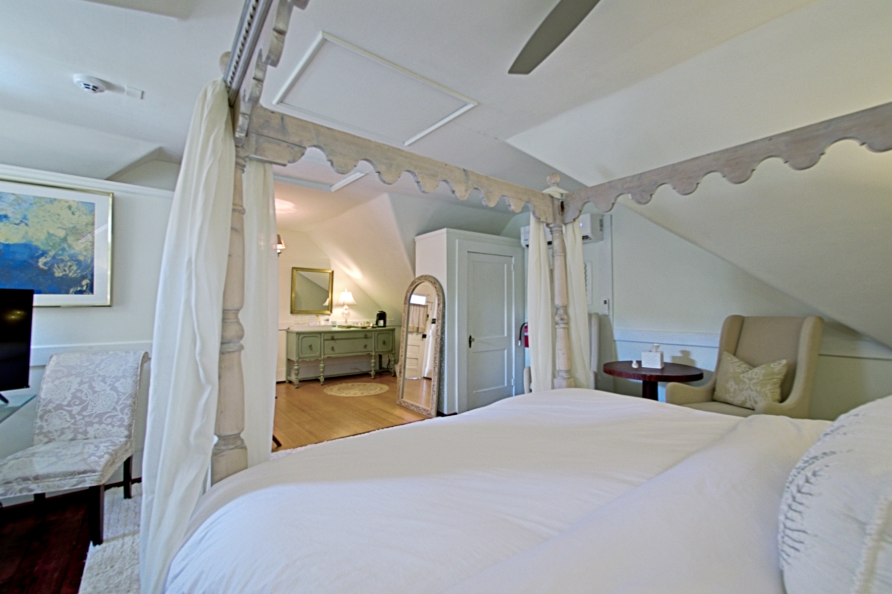 A symmetrical, wide-angle shot of an attic-style bedroom featuring a white four-poster canopy bed with flowing white curtains and a decorative scalloped frame. The room has sloped white ceilings and dark wood floors, with a beige armchair on the left and a matching chaise lounge on the right. A plush white rug sits at the foot of the bed, and a ceiling fan is visible above.