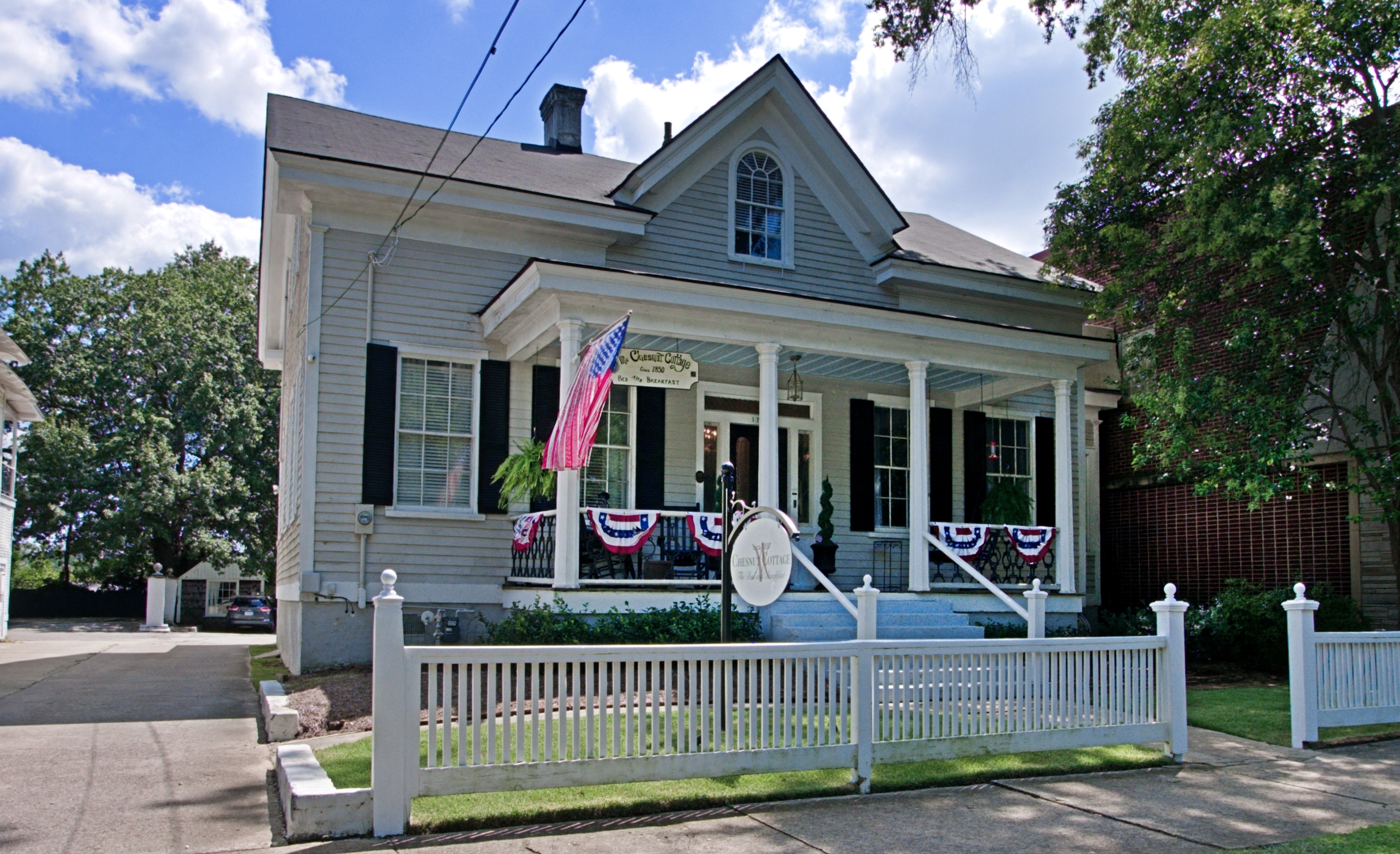 An exterior wide-angle shot of a light grey one-and-a-half-story house with a dark roof and black shutters. The home features a covered front porch supported by white columns, decorated with patriotic red, white, and blue bunting and an American flag. A white picket fence runs along the front of the property, and a white sign is posted in the yard.