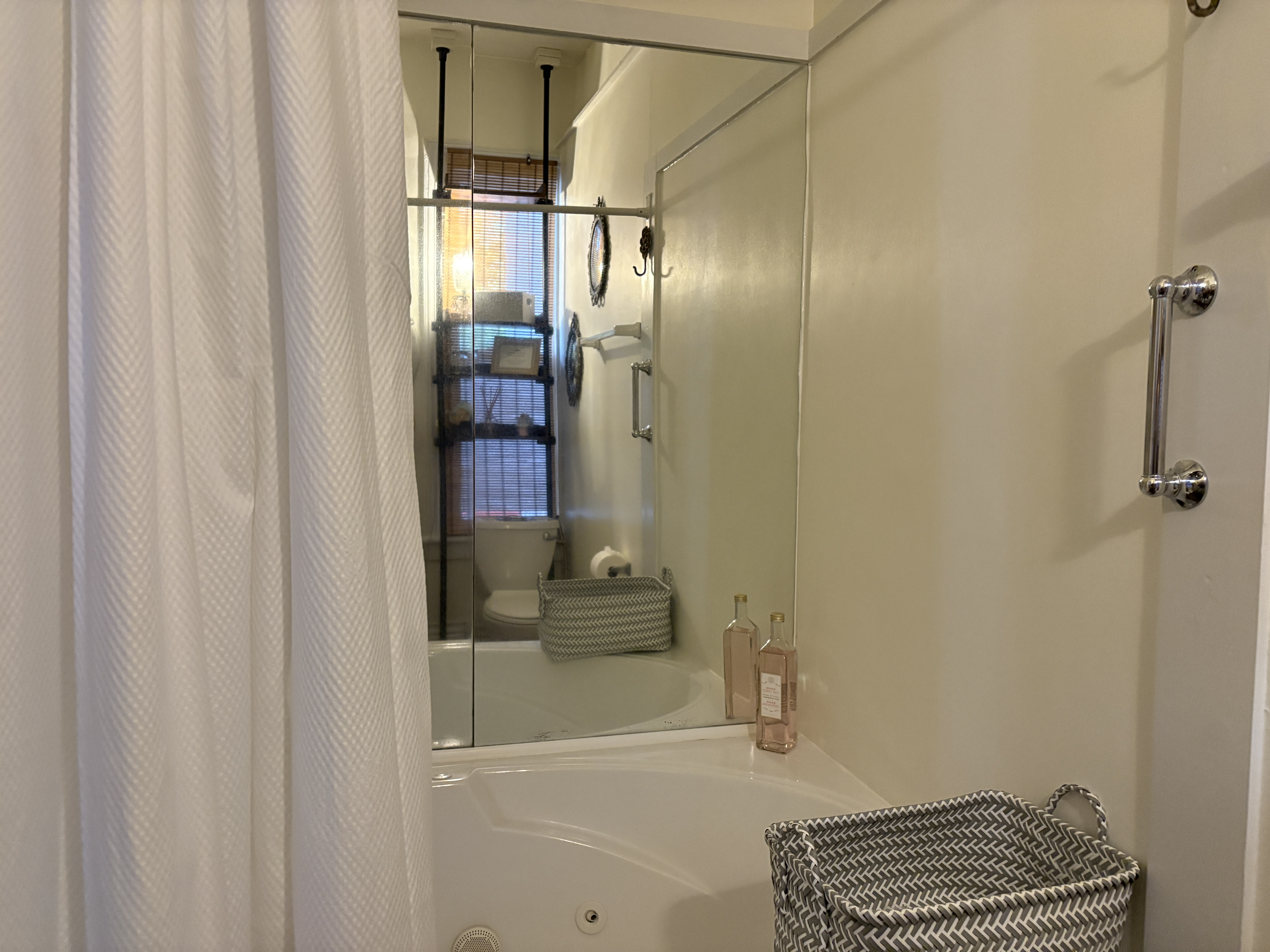 A close-up shot of a bathroom featuring a large white soaking tub and a white shower curtain. A large mirror on the back wall reflects a shelving unit holding various toiletries, while two glass bottles and a grey-and-white striped basket sit on the edge of the tub in the foreground.