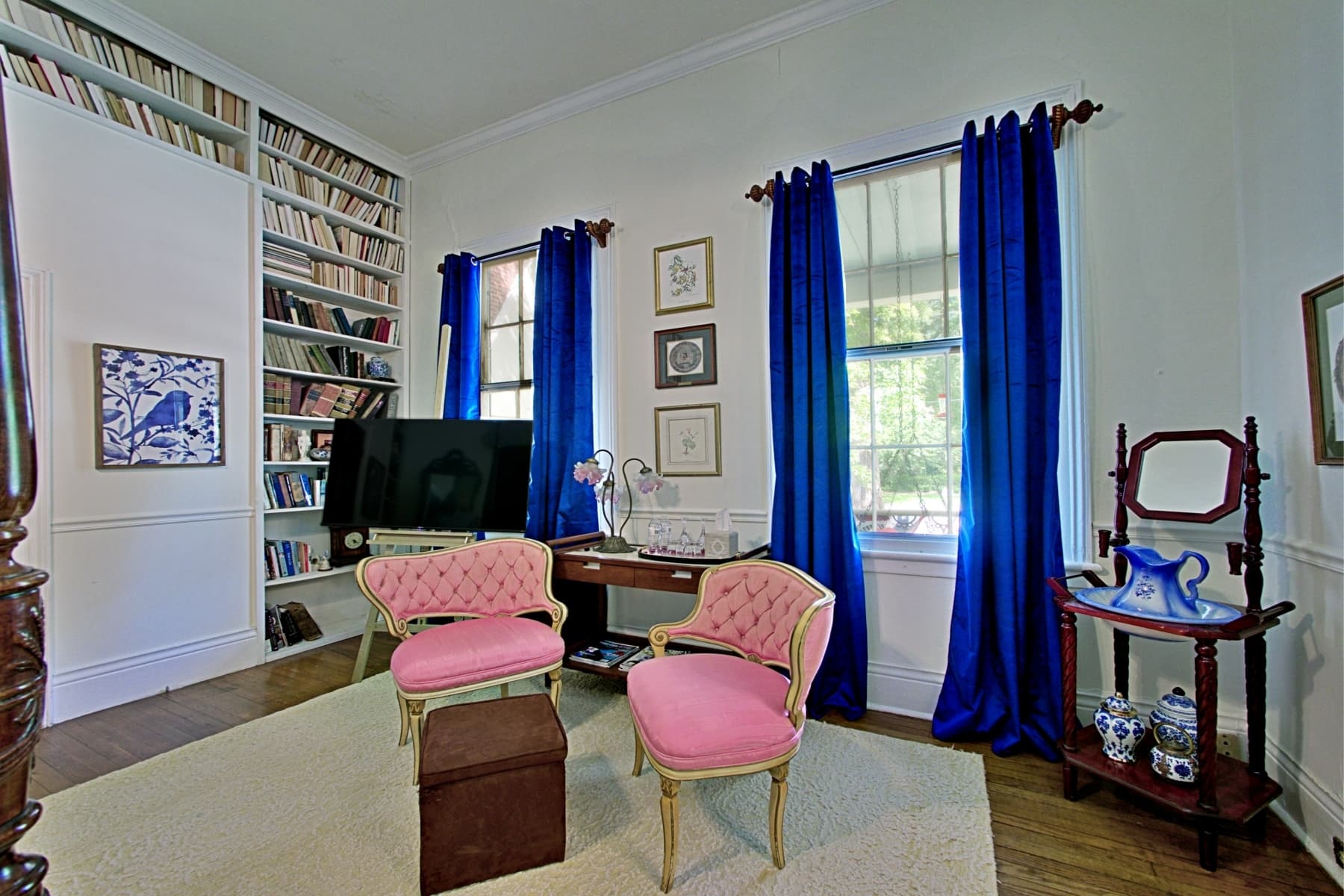 A brightly lit sitting area featuring two pink tufted armchairs arranged around a dark wood desk and a small brown ottoman. The room includes floor-to-ceiling built-in bookshelves, two windows framed by vibrant blue curtains, and a wooden washstand with a blue and white pitcher.