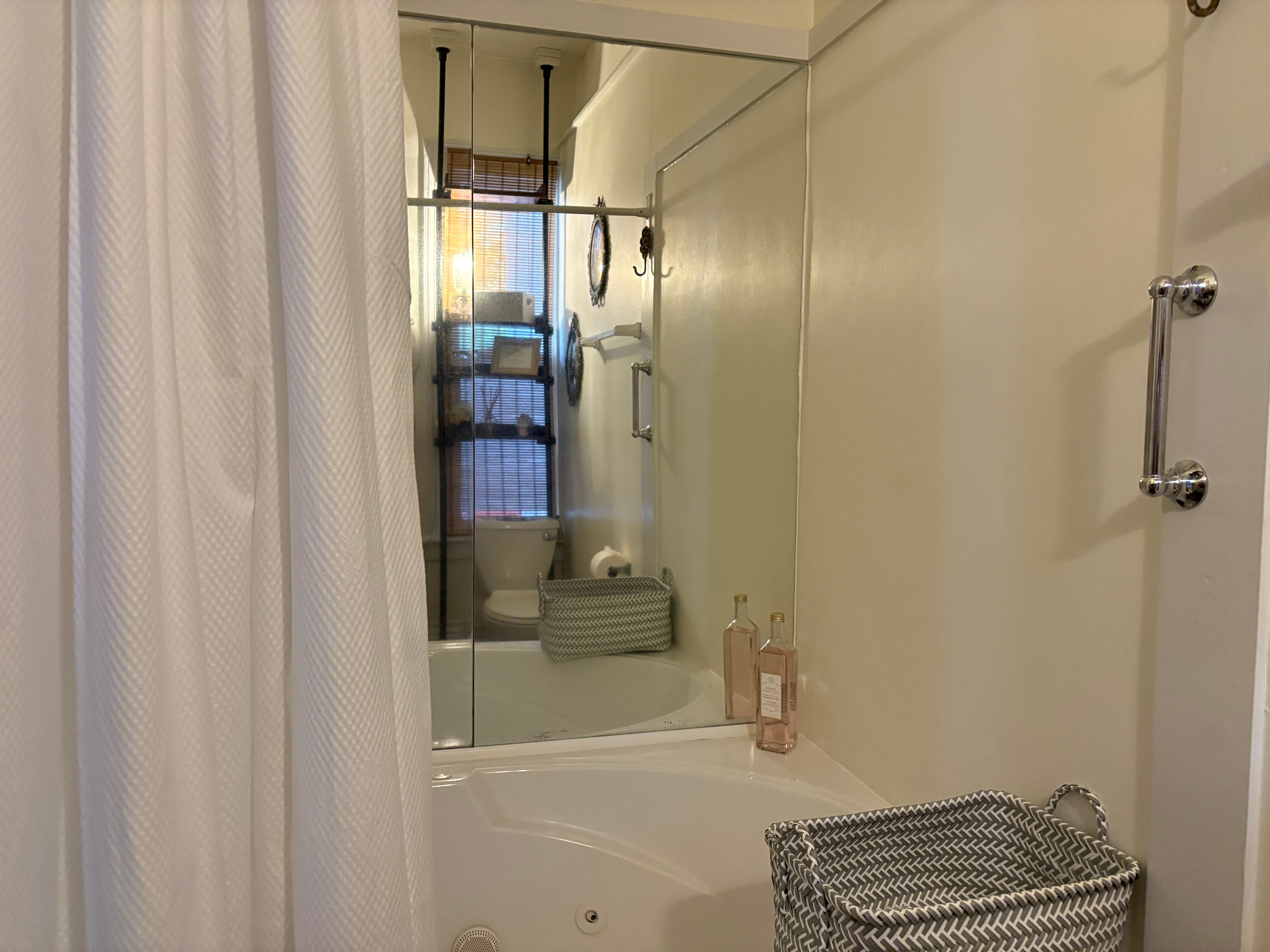 A high-angle shot of a white alcove bathtub with a white shower curtain pulled to one side. A grey and white herringbone-patterned basket sits on the corner of the tub, and a large mirror on the back wall reflects a toilet and a dark metal storage rack.