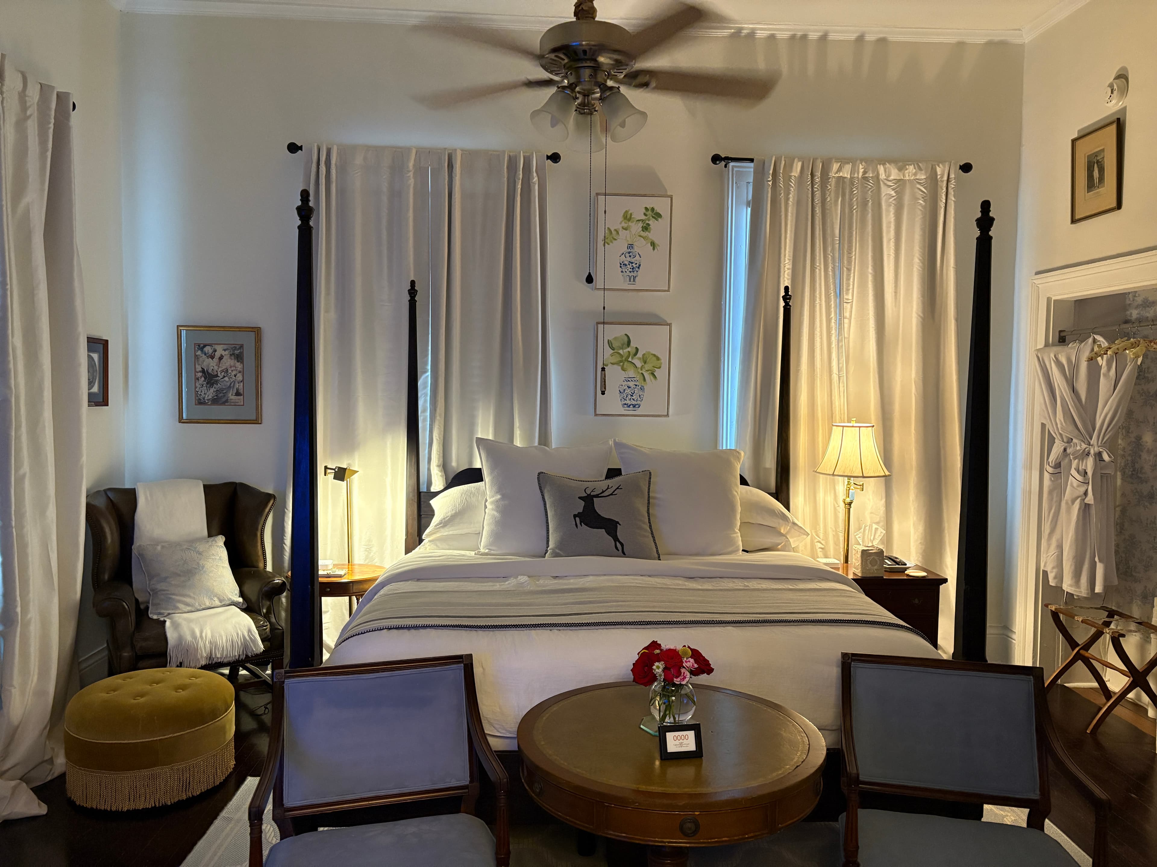 A symmetrical shot of a bedroom featuring a dark four-poster bed with white linens and a grey throw. In the foreground, two small blue chairs flank a round wooden table with a vase of red roses, while a black leather wingback chair with a yellow ottoman sits to the left. The room is decorated with framed botanical prints and two windows draped in long white curtains.