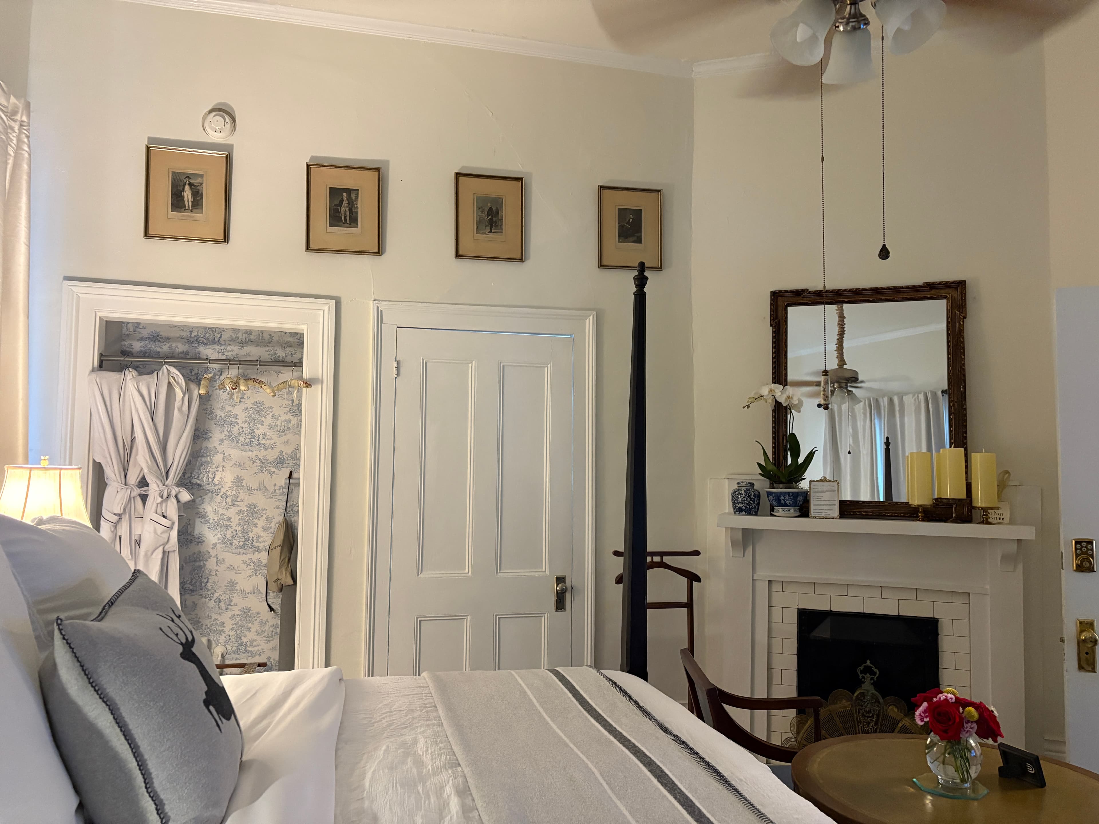 An elegant bedroom featuring a dark wood four-poster bed with crisp white linens and a grey throw blanket. In the foreground, two light blue chairs face a round wooden table topped with a small vase of red flowers, while a dark leather wingback chair with a yellow ottoman sits to the left. The room is accented with long white curtains framing the windows and several framed botanical prints on the walls.