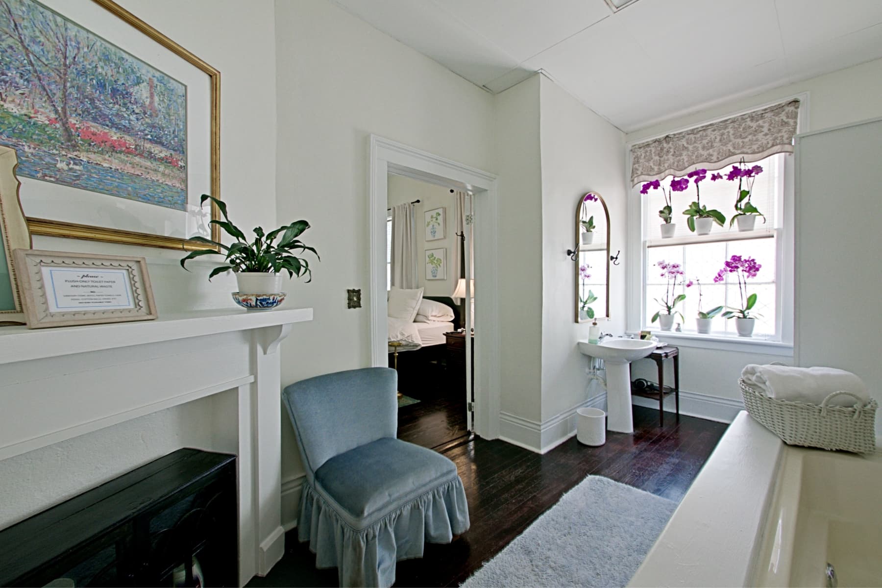 A bright suite featuring dark wood floors and white walls, with a white pedestal sink positioned under a window adorned with purple orchids. The room includes a large white mantel with a framed landscape painting, a light blue velvet vanity chair, and a white bathtub partially visible in the foreground.
