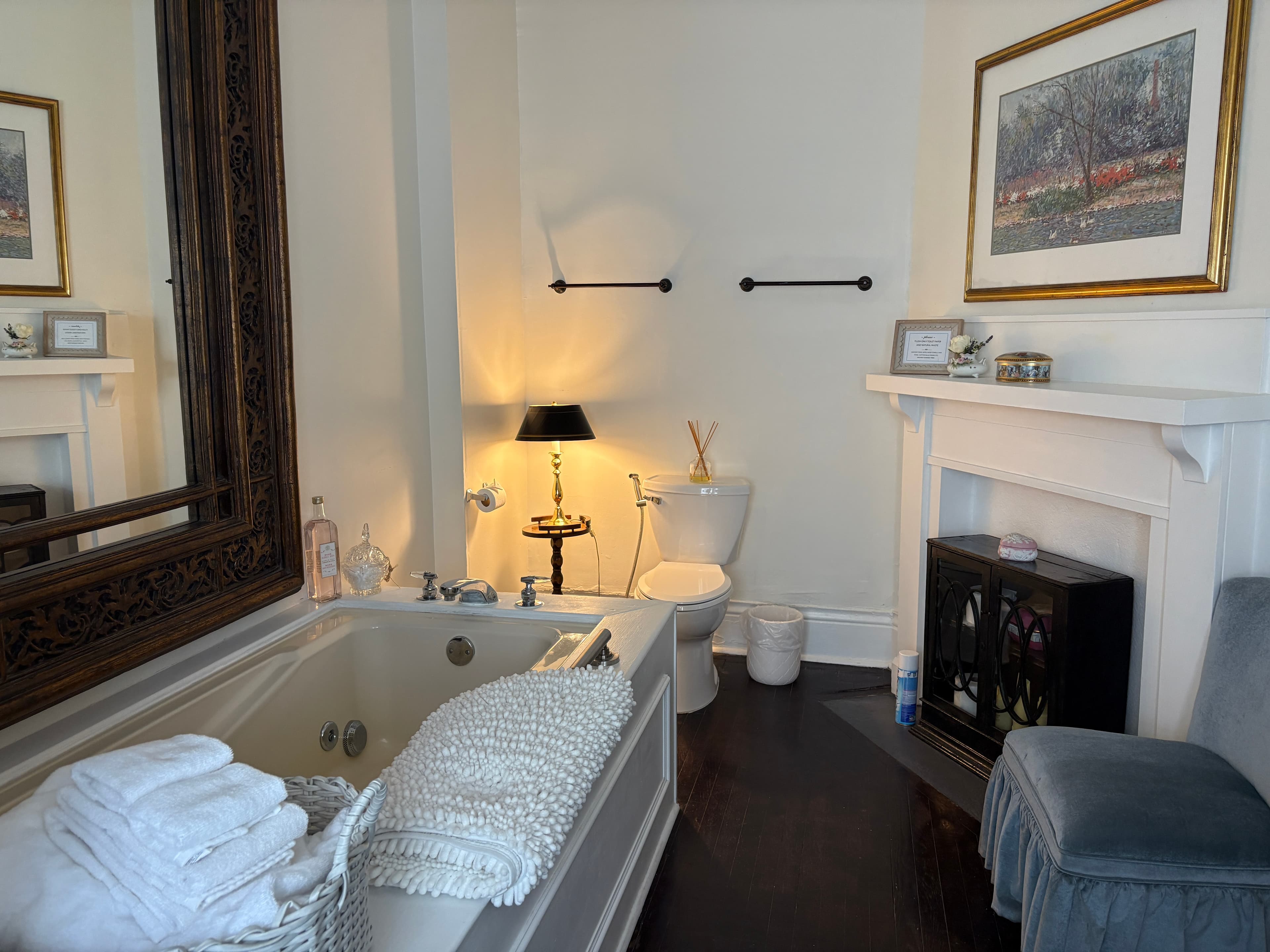 A bright, elegant suite featuring dark wood floors and white walls, with a white pedestal sink positioned under a window adorned with vibrant purple orchids. The room includes a large white mantel with a framed landscape painting, a light blue velvet vanity chair, and a white bathtub partially visible in the foreground.