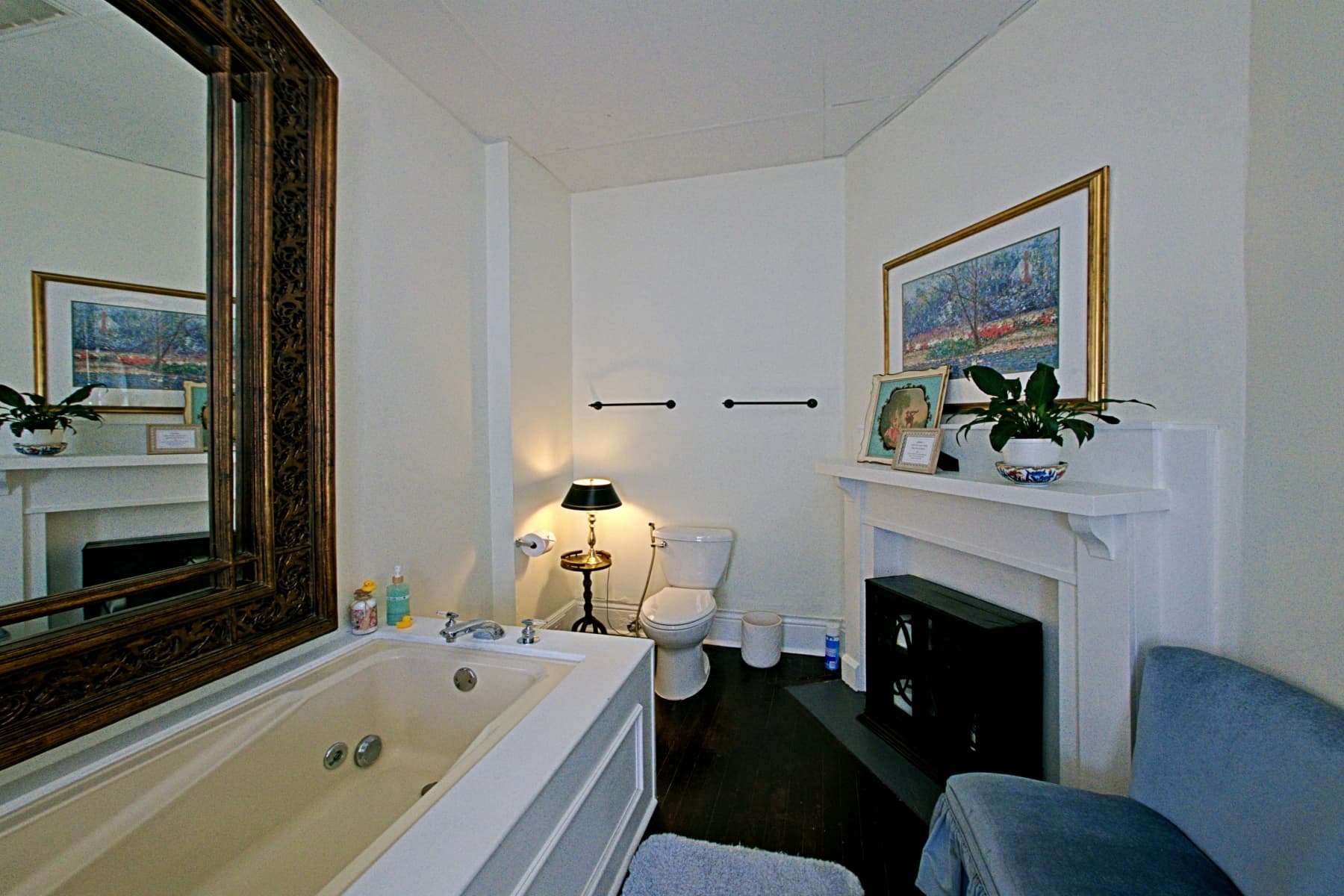 A bright, elegant bathroom featuring dark wood floors and white walls, with a white pedestal sink positioned under a window adorned with vibrant purple orchids. The room includes a large white mantel with a framed landscape painting, a light blue velvet vanity chair, and a white bathtub partially visible in the foreground.