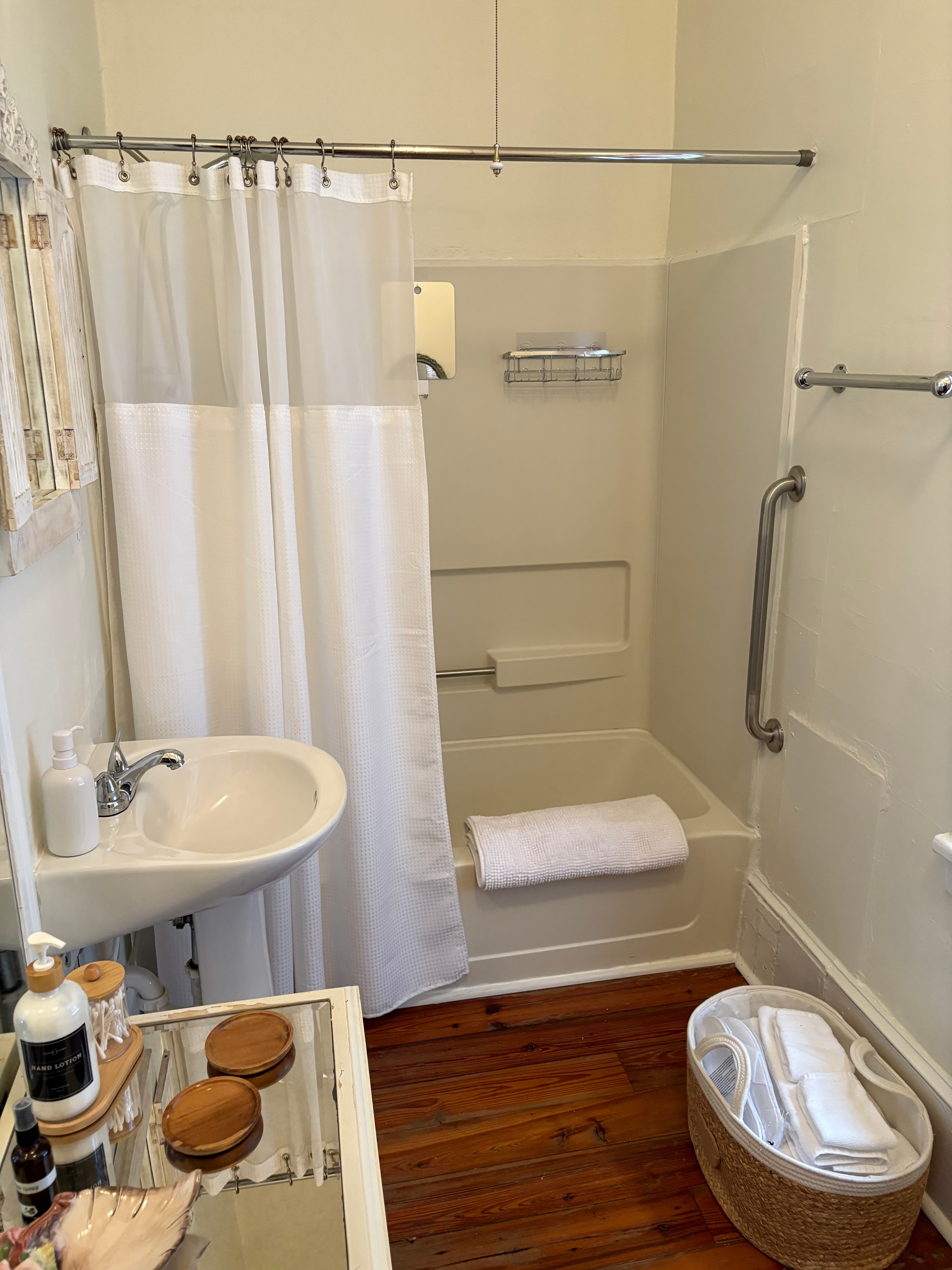 A brightly lit, narrow bathroom featuring a white pedestal sink and a matching white bathtub and shower with a white curtain. The space is accented with natural textures, including a bamboo-style window shade and a white woven basket on the dark wood flooring.