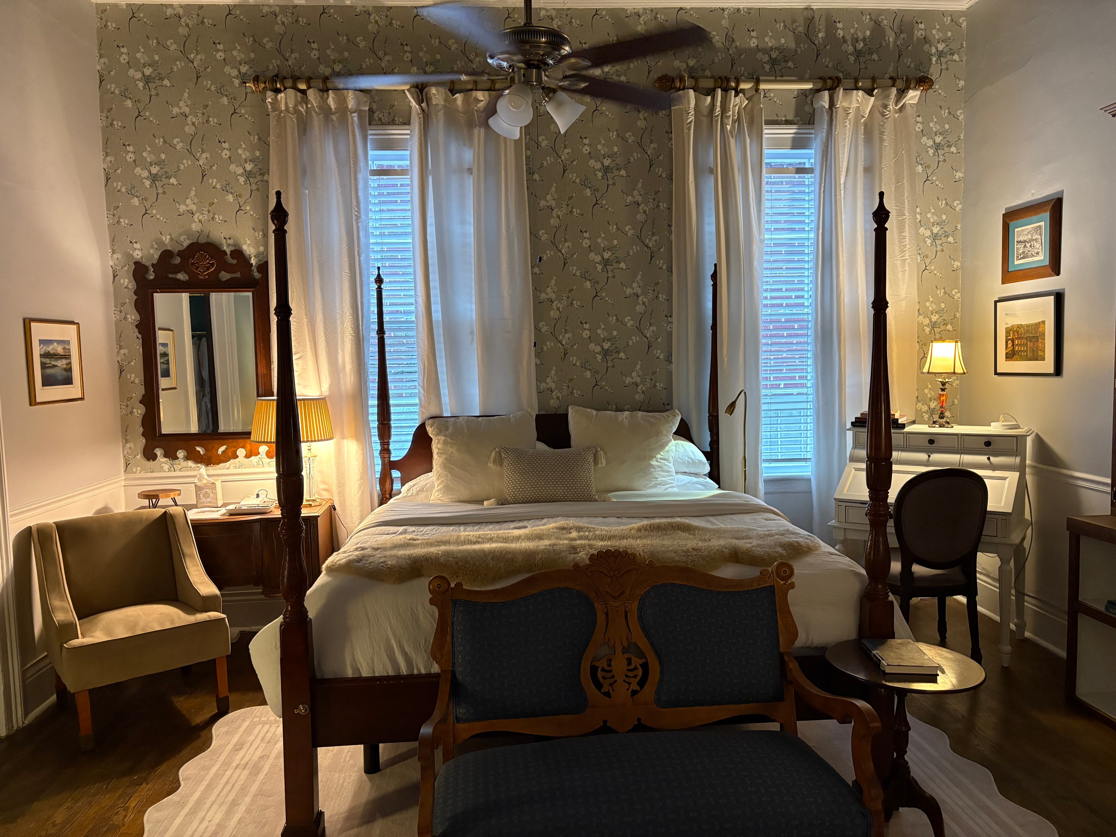 An elegant bedroom featuring a dark wood four-poster bed with a white duvet and a brown faux-fur throw. In the foreground, an ornate blue-upholstered bench sits at the foot of the bed, while a small round side table with a lamp is positioned to the left. A doorway leads to another room with dark teal walls, and a large mirror with a decorative frame is visible on the wall to the right.