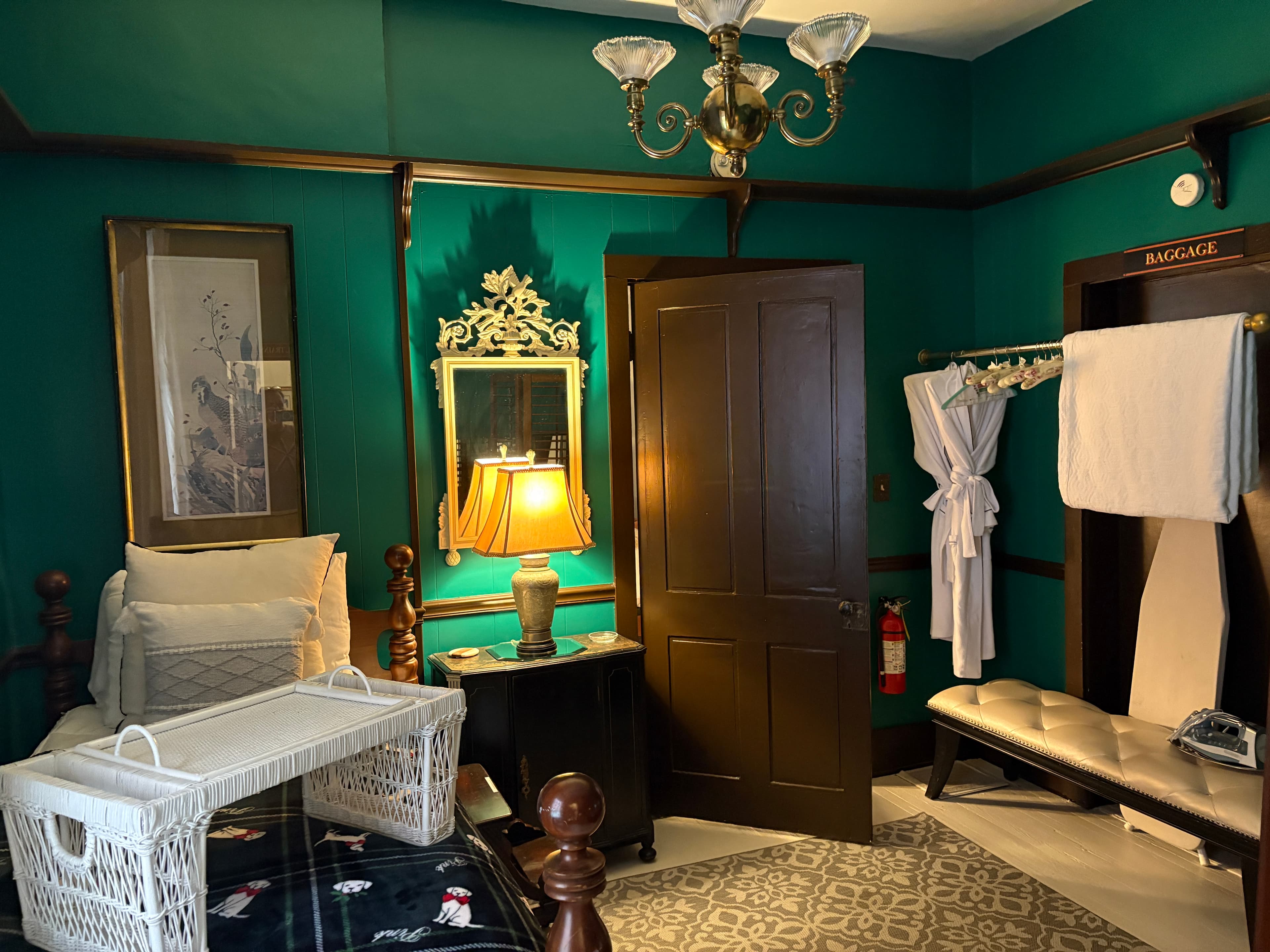 A bedroom with deep teal walls and dark wood accents features a bed with white pillows and a white wicker breakfast tray. To the right of a closed wooden door, a white bathrobe and towels hang on a rack next to a padded bench and an ironing board. An ornate gold mirror and a lamp with a warm glow are positioned on a small black nightstand to the left of the door.