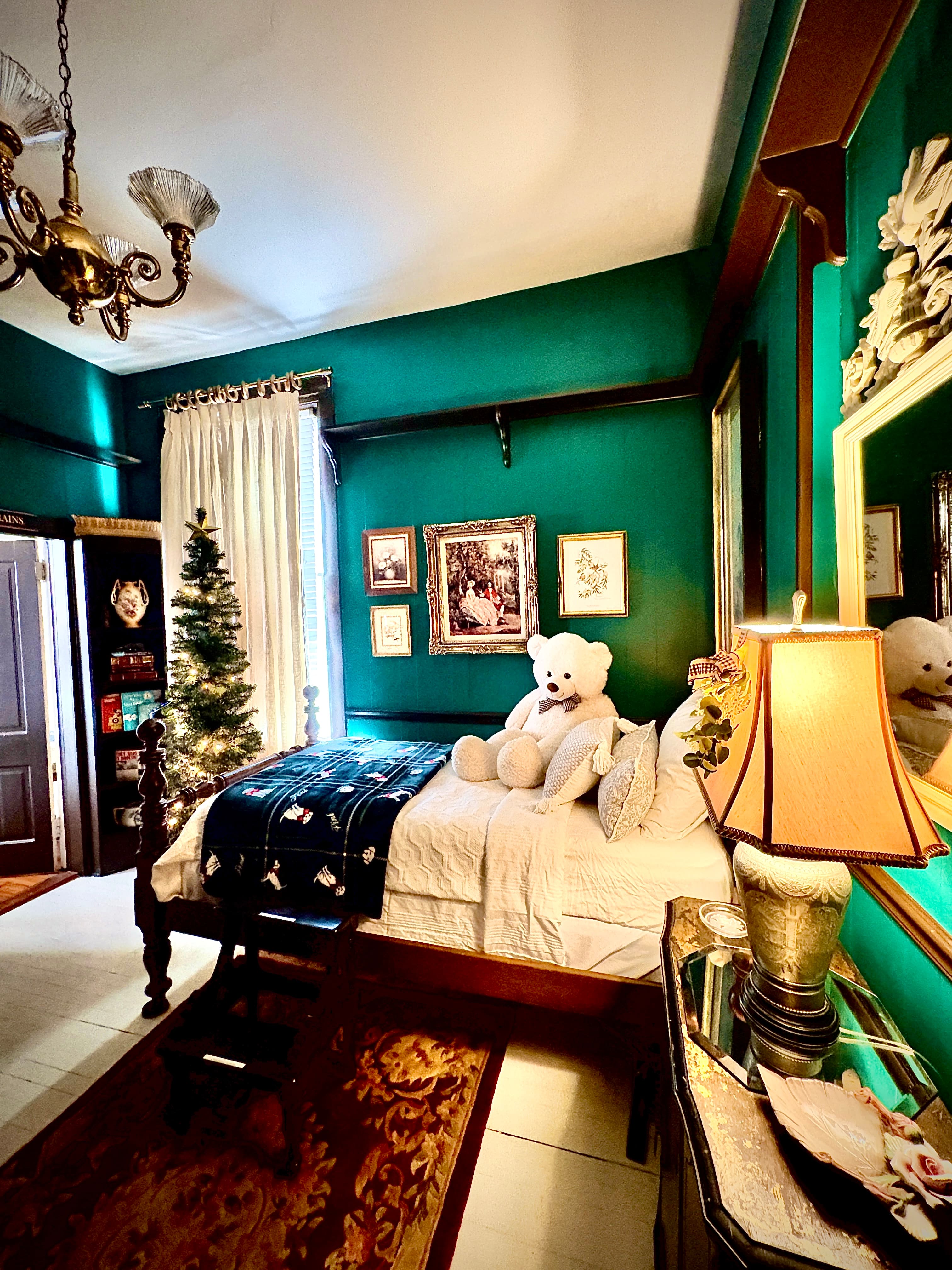 A cozy bedroom with vibrant teal walls and dark wood trim features a bed with white linens and a large white teddy bear sitting against the pillows. To the left of the bed stands a lit Christmas tree in front of a window with white curtains, and a variety of framed pictures are arranged on the wall above the bed.