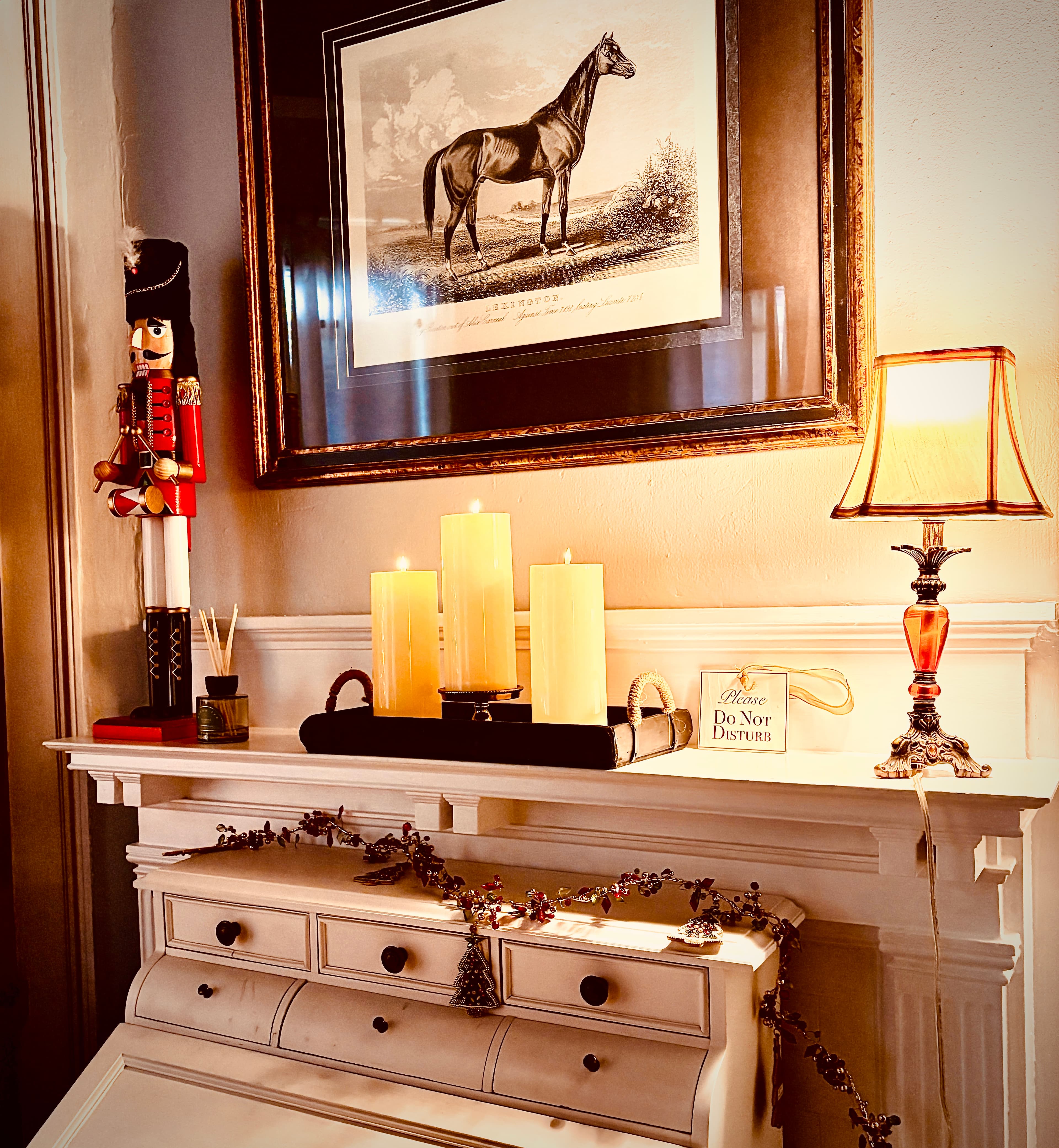 A warm, close-up shot of a white mantel decorated for the holidays. On top of the mantel sits a black tray with three lit pillar candles, a traditional nutcracker figurine, and a small lamp with an amber base. Above the mantel hangs a large framed illustration of a horse, and below it, a white vintage-style secretary desk is adorned with a festive garland.