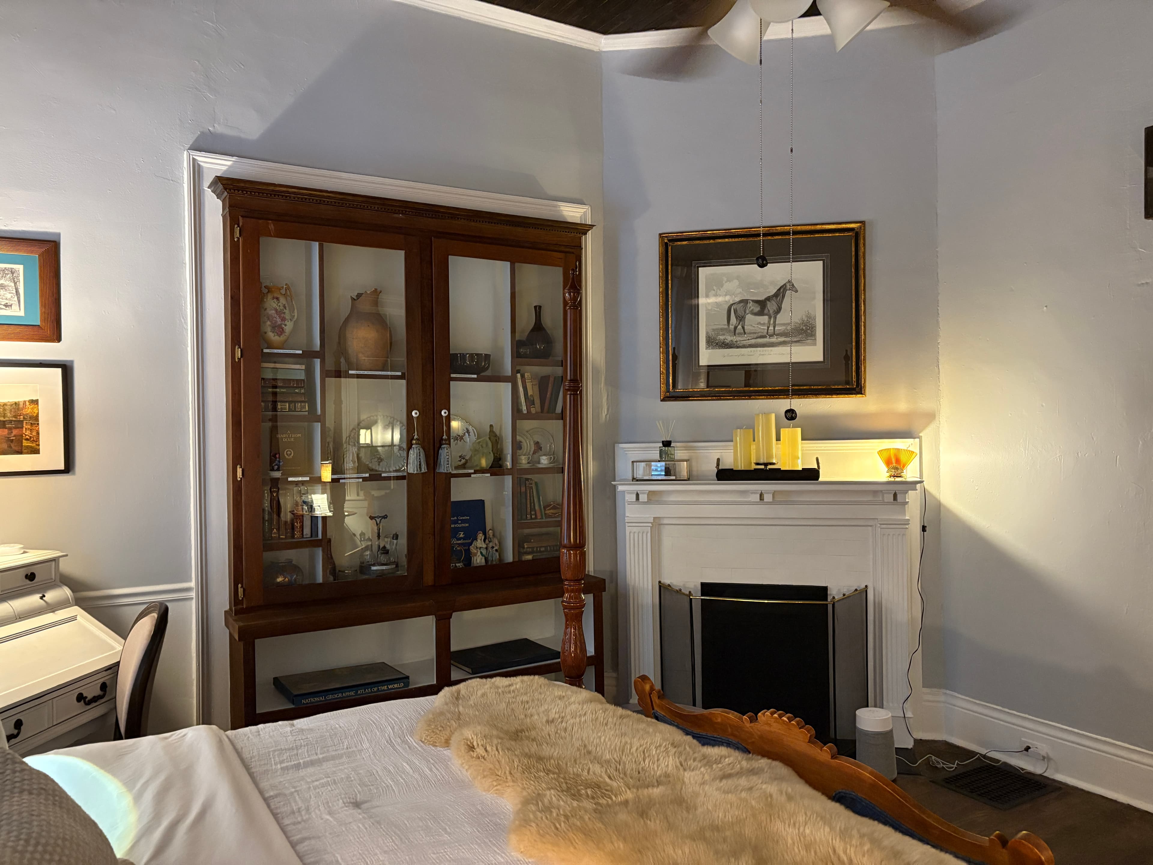 A wide-angle view of a traditional bedroom featuring a dark wood four-poster bed with a sheepskin throw and an ornate blue-upholstered bench. The room includes a large wooden display cabinet filled with collectibles, a white fireplace mantel with lit candles and a horse illustration above it, and a white secretary desk in the corner.
