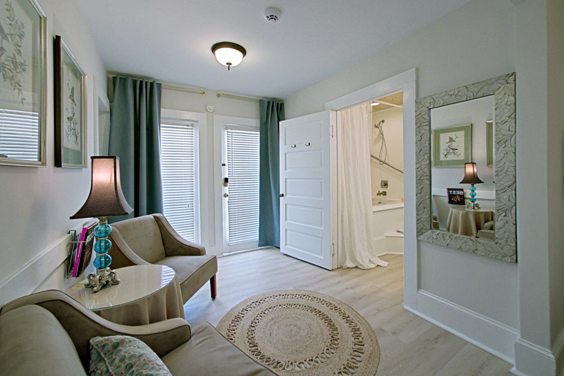 A brightly lit interior featuring a sitting area with two beige armchairs and a small round table in the foreground. In the background, a wide doorway leads into a bathroom area where a white shower curtain is visible. The room is decorated with a round jute rug, a tall mirror with a textured frame, and light-colored wood flooring.