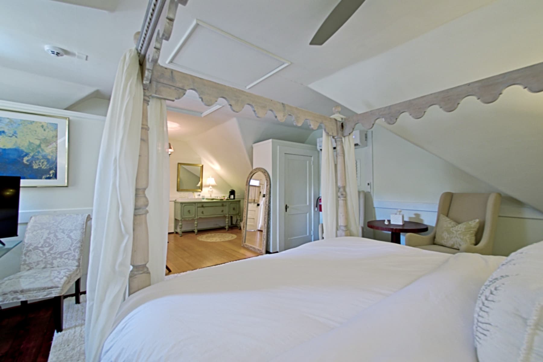 A symmetrical, wide-angle shot of an attic-style bedroom featuring a white four-poster canopy bed with flowing white curtains and a decorative scalloped frame. The room has sloped white ceilings and dark wood floors, with a beige armchair on the left and a matching chaise lounge on the right. A plush white rug sits at the foot of the bed, and a ceiling fan is visible above.