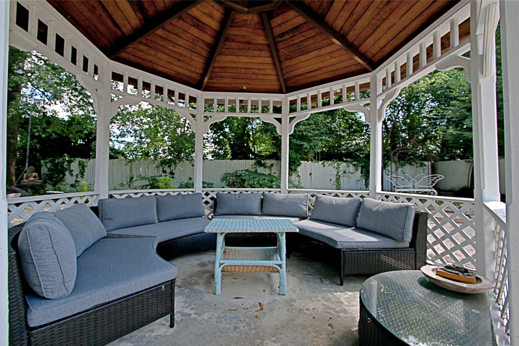 An outdoor white wooden gazebo with a conical wood-plank ceiling and lattice railings. The interior features a large curved wicker sectional sofa with grey cushions surrounding a small light-blue table, overlooking a lush green garden.