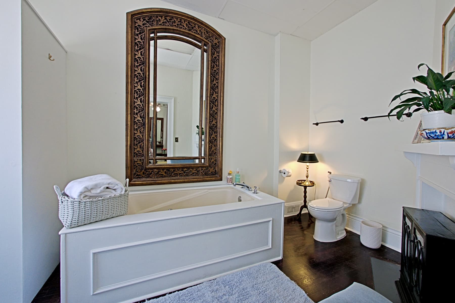 A spacious white bathroom featuring a large rectangular soaking tub with an ornate, arched wood-framed mirror mounted above it. To the right is a white toilet, a small side table with a lamp, and dark flooring.