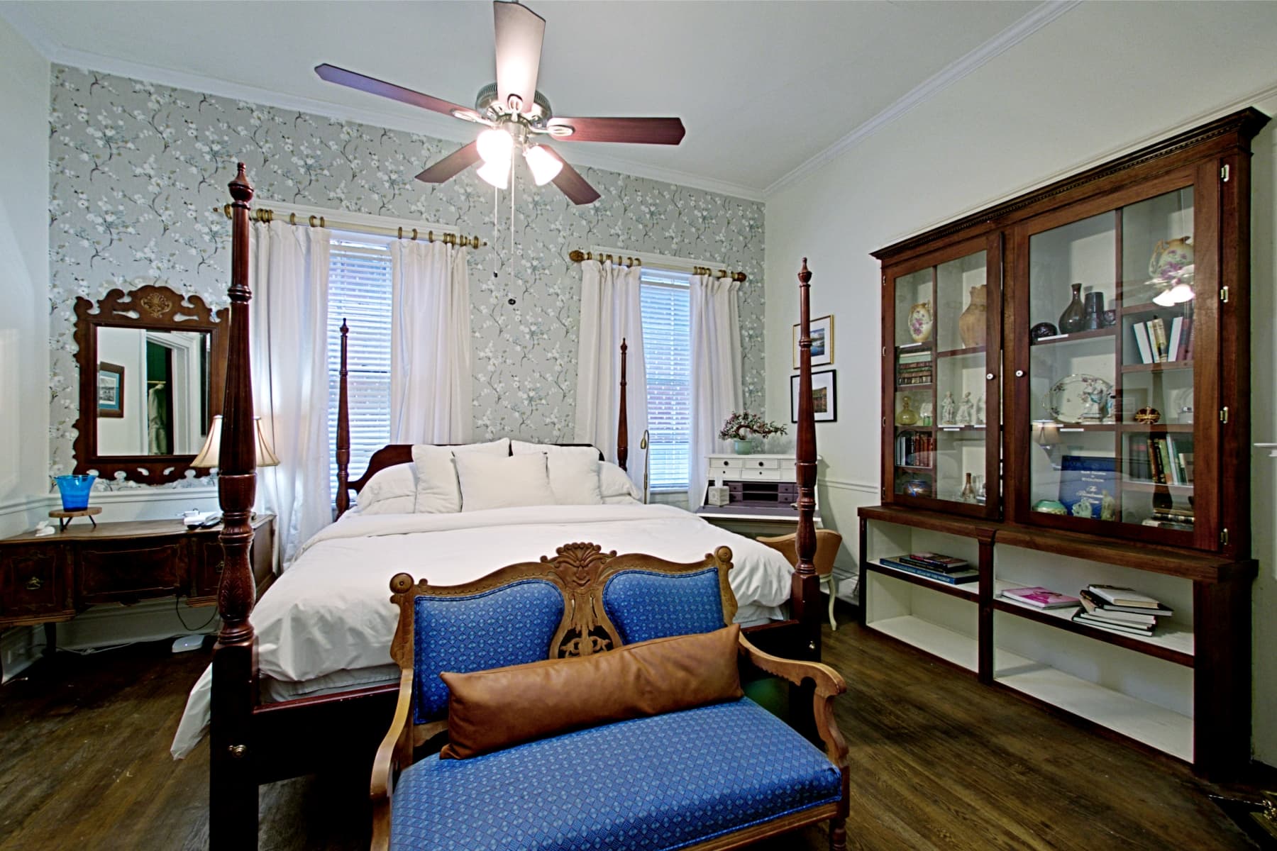 A traditional bedroom featuring a dark wood four-poster bed with white linens and a blue settee at the foot. The room includes a patterned accent wall, two windows with white curtains, a lit ceiling fan, and a large wooden display cabinet on the right.