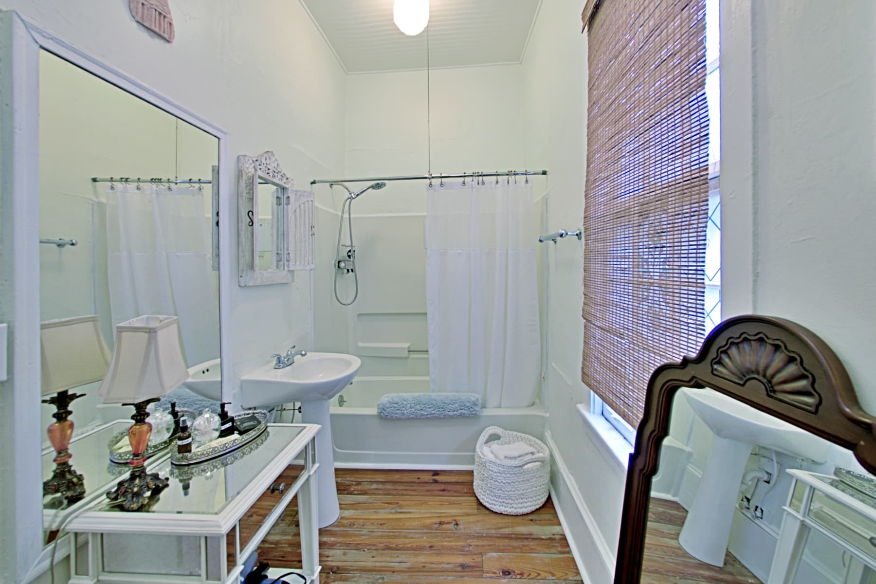 A white-walled bathroom featuring a pedestal sink, a shower-tub combination with a white curtain, and wood-plank flooring. The room includes a mirrored vanity table with a lamp on the left and a window with a bamboo blind on the right.