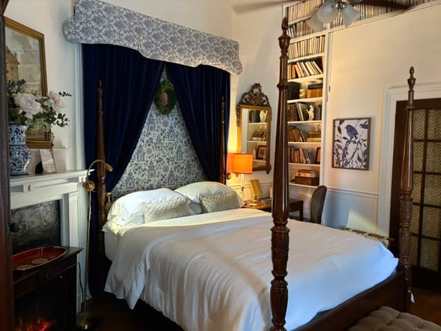 A traditional, cozy bedroom featuring a four-poster wooden bed with white linens, set against a backdrop of ornate blue and white patterned wallpaper and deep blue velvet curtains hanging from a matching valance. A tall white bookshelf filled with books, an ornate gold mirror, and a small desk with an orange lamp sit to the right of the bed.