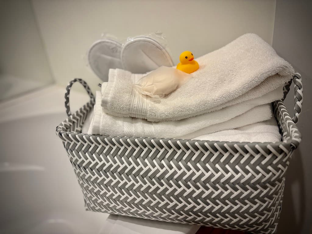 A grey and white herringbone-patterned basket filled with folded white towels sits on the edge of a white bathtub. Tucked into the top of the basket are a pair of white slippers, a bar of soap, and a small yellow rubber duck.