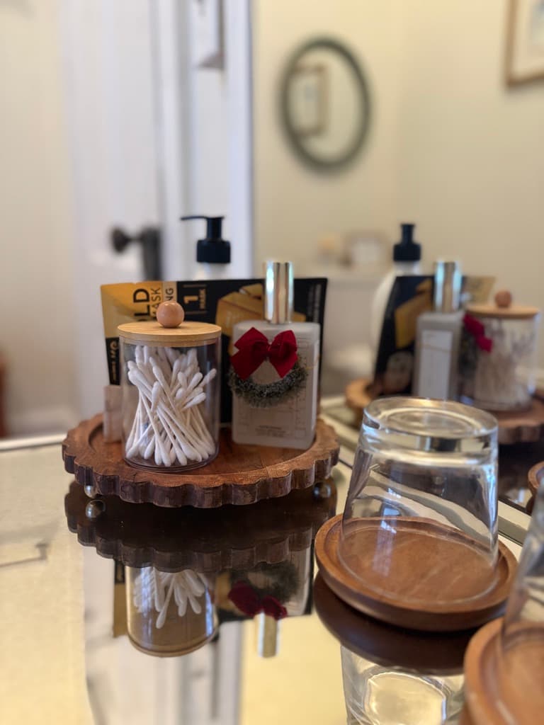A close-up shot of a mirrored surface reflecting several bathroom amenities arranged on a circular wooden tray. The items include a glass jar of cotton swabs, a bottle of lotion adorned with a small festive red bow and wreath, and a pump bottle. An upside-down drinking glass on a wooden coaster sits in the foreground.