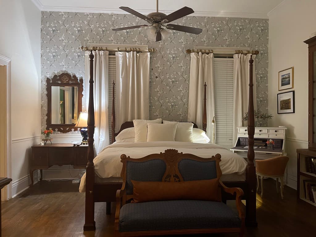 A wide-angle view of a traditional bedroom featuring a dark wood four-poster bed with a blue and white patterned quilt. The room includes a fireplace with a white mantel, a large wooden armoire with a mirrored door, a ceiling fan, and built-in bookshelves that extend to the ceiling.