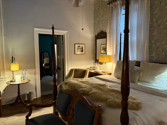 An elegant bedroom featuring a dark wood four-poster bed with a white duvet and a brown faux-fur throw. In the foreground, an ornate blue-upholstered bench sits at the foot of the bed, while a small round side table with a lamp is positioned to the left. A doorway leads to another room with dark teal walls, and a large mirror with a decorative frame is visible on the wall to the right.