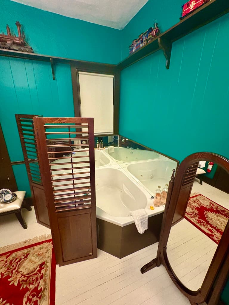 A wide-angle view of a traditional bedroom featuring a dark wood four-poster bed with a blue and white patterned quilt. The room includes a fireplace with a white mantel, a large wooden armoire with a mirrored door, a ceiling fan, and built-in bookshelves that extend to the ceiling.