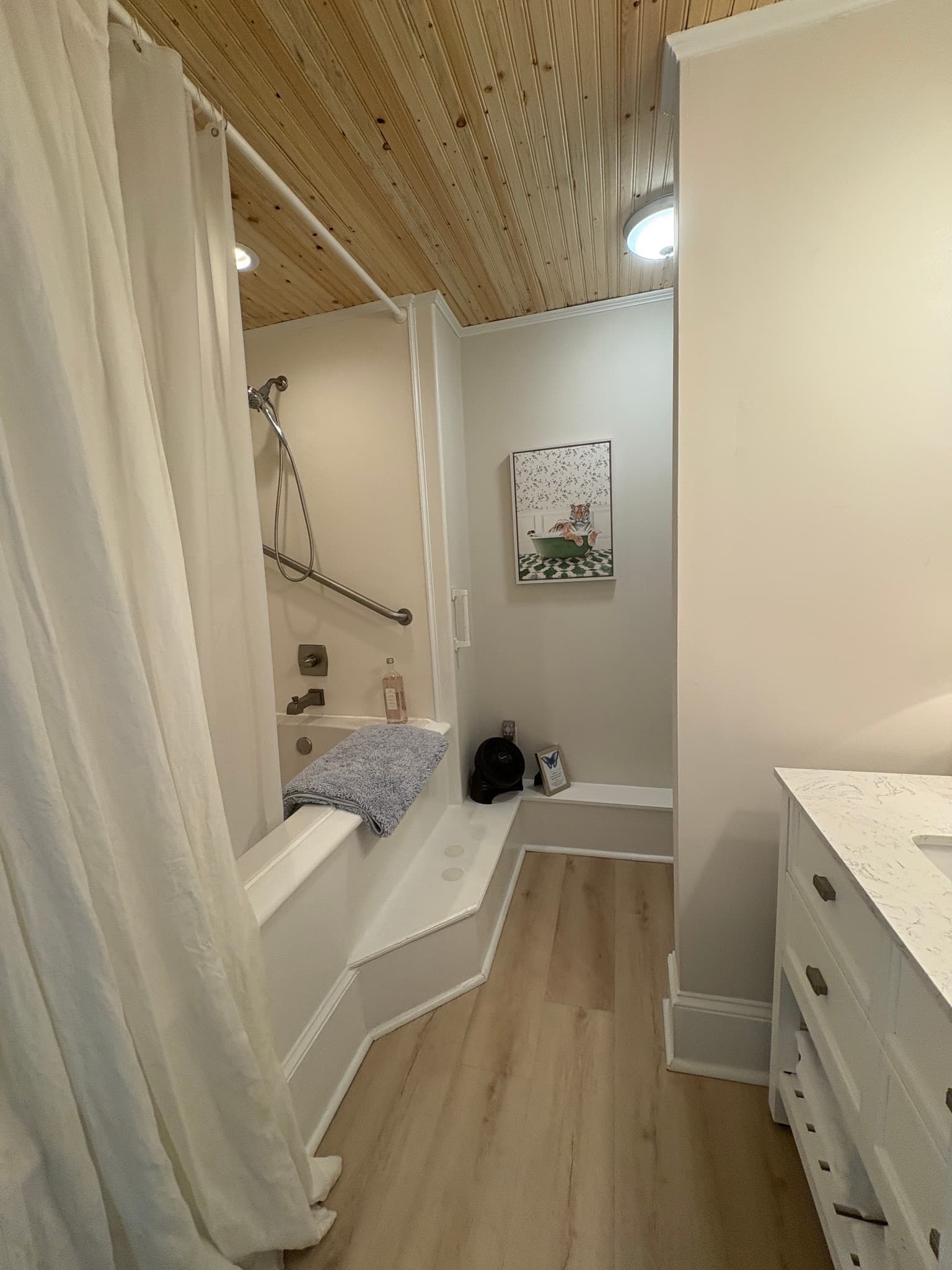 A brightly lit bathroom with light wood-look flooring and a light-colored wood-plank ceiling. On the left, a white bathtub with a white shower curtain is partially visible, while a white vanity with a marble top is on the right. The back wall features a small framed picture above a white ledge.