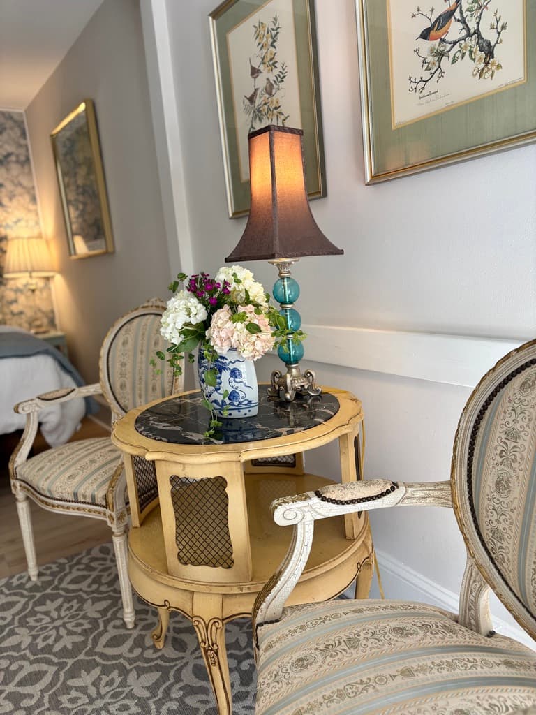 A close-up shot of an elegant sitting area featuring two ornate cream-colored armchairs with striped upholstery. Between them sits a small, two-tier gold side table with a black marble top, holding a blue and white floral vase with fresh flowers and a lamp featuring a blue glass base and a dark shade. Two framed bird illustrations hang on the wall above the seating.