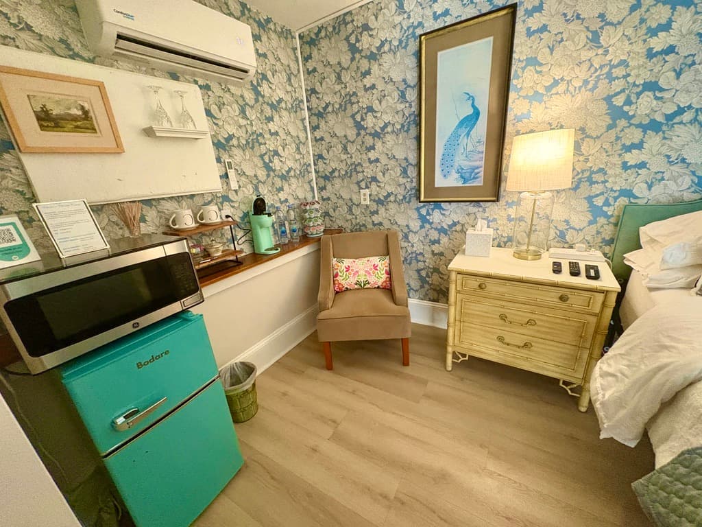 A wide-angle view of a traditional bedroom featuring a dark wood four-poster bed with a blue and white patterned quilt. The room includes a fireplace with a white mantel, a large wooden armoire with a mirrored door, a ceiling fan, and built-in bookshelves that extend to the ceiling.