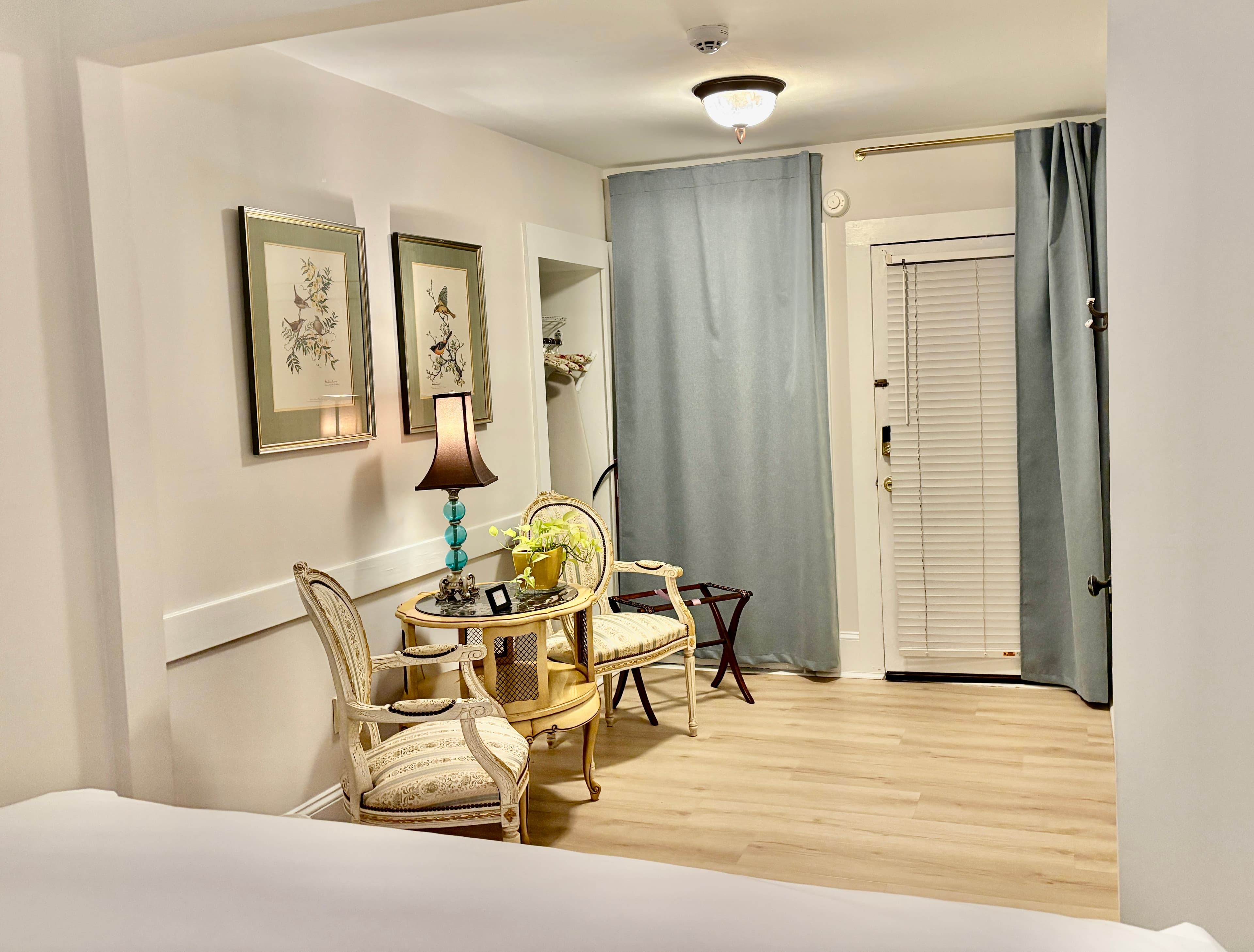 A wide-angle shot of a cozy sitting area with light wood flooring, featuring two beige armchairs and a small round table with a blue-glass lamp. A wide doorway leads to a bright bathroom area where a white shower curtain and a large wall mirror are visible. The room is accented with a round jute rug and framed botanical prints on the wall.
