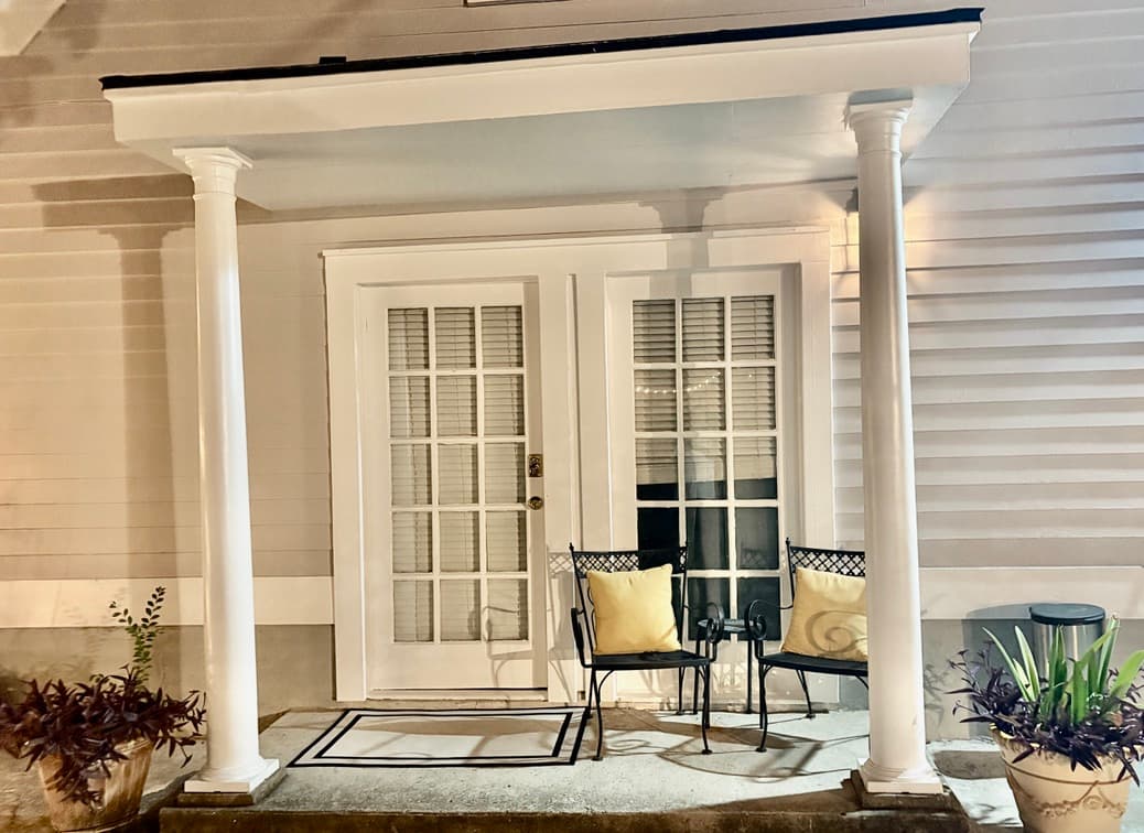 A well-lit outdoor patio at night featuring two dark metal chairs with beige cushions positioned in front of white-trimmed French doors. The covered area is supported by two white architectural columns and includes a small patterned rug on the floor and potted plants on either side.