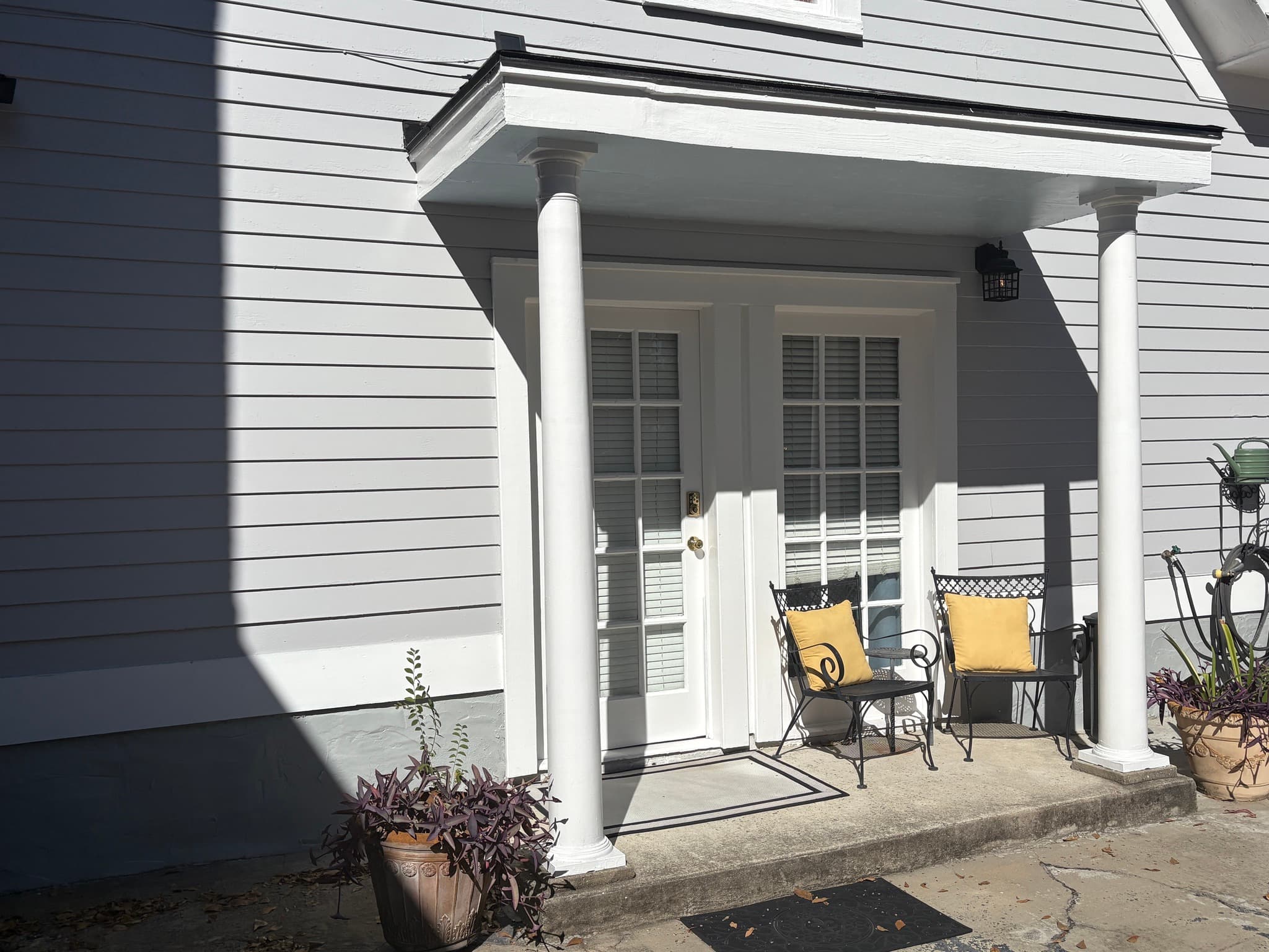 A daylight wide-angle view of a grey siding exterior featuring a covered porch with white architectural columns. Two dark metal chairs with yellow cushions sit in front of white-trimmed French doors, flanked by potted plants on a concrete patio.