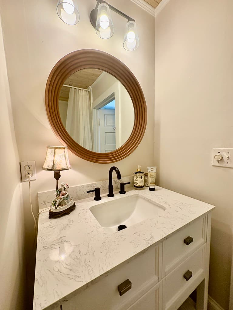 A white bathroom vanity with a marble countertop and a black faucet is situated beneath a large, round wooden mirror. The vanity holds a small lamp with an ornate base and a bottle of soap, while three modern light fixtures are mounted on the wall above the mirror.