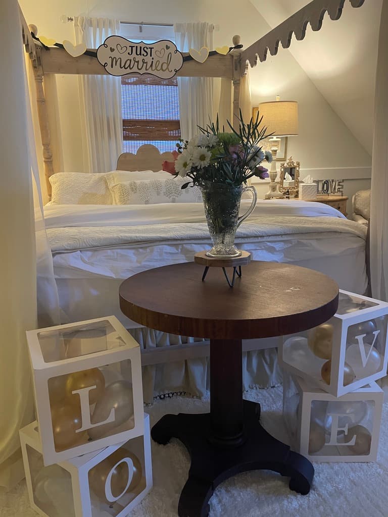 A romantic setup in an attic-style bedroom features a dark wood pedestal table with a clear glass pitcher of flowers. In the background, a white four-poster canopy bed is decorated with a "Just Married" sign and heart-shaped accents, while white cubes that spell out "LOVE" are stacked on the floor in the foreground.