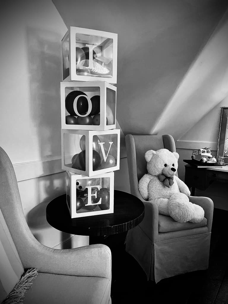 A black-and-white photograph showing a corner of an attic-style bedroom with sloped ceilings. A stack of four white cubes spelling out "LOVE" sits on a dark round table, while a large white teddy bear rests in a nearby beige wingback chair.