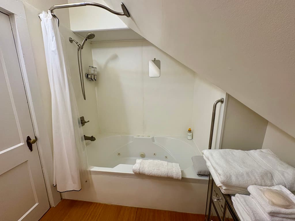 A bathroom located under a sloped attic ceiling features a large white soaking tub with chrome fixtures and a white shower curtain. To the right, a mirrored vanity holds a stack of fresh white towels and spa slippers, while a wooden floor runs throughout the space.
