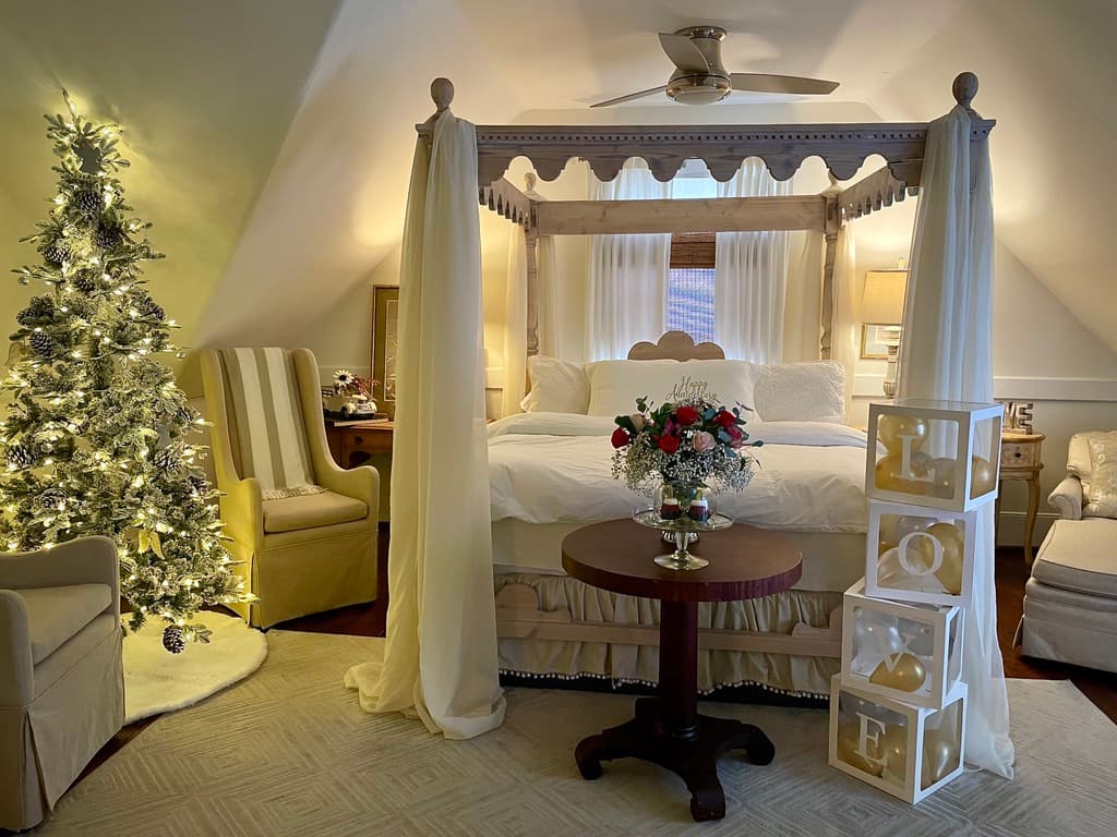 A symmetrical, wide-angle shot of an attic-style bedroom featuring a white four-poster canopy bed with flowing white curtains and a decorative scalloped frame. The room has sloped white ceilings and dark wood floors, with a beige armchair on the left and a matching chaise lounge on the right. A plush white rug sits at the foot of the bed, and a ceiling fan is visible above.