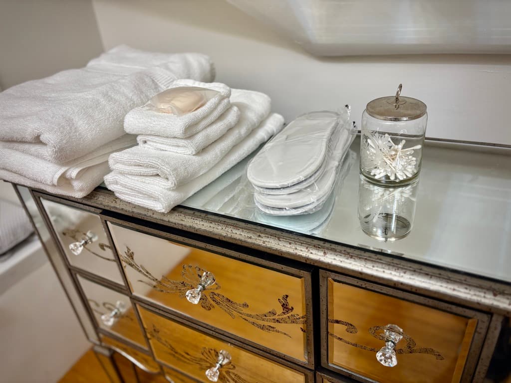 A close-up shot of a mirrored dresser with ornate floral etchings on the drawers. The glass top is neatly arranged with guest amenities, including a stack of white folded towels, a bar of soap, a pair of white spa slippers, and a glass jar filled with cotton swabs.