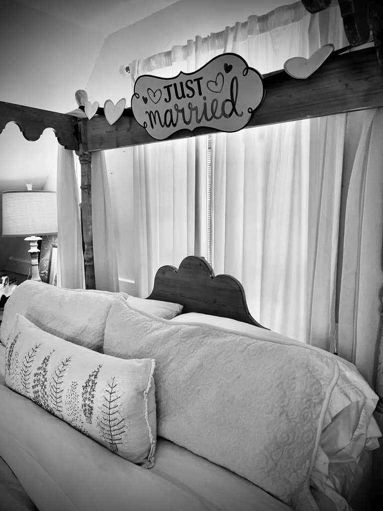 A black-and-white, close-up photograph of a bed's headboard area, featuring a decorative "Just Married" sign adorned with small hearts hanging from the top crossbar of a four-poster bed. The bed is neatly made with several white pillows, including a long lumbar pillow with a botanical leaf pattern, set against white window curtains in the background.
