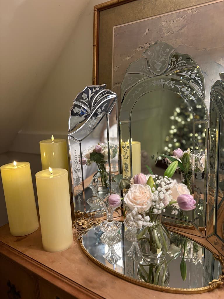 A close-up shot of an elegant vanity area featuring three lit pillar candles on the left. In the center, a bouquet of pink roses and white baby's breath is displayed in a glass vase, resting on an oval mirrored tray. The scene is reflected in an ornate three-panel mirror, which also captures the soft glow of a Christmas tree in the background.