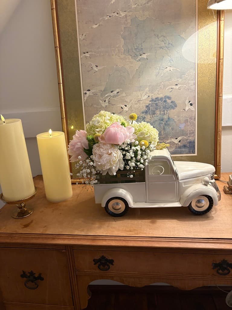 A decorative wooden dresser top features two lit pillar candles on the left and a white vintage-style ceramic truck on the right. The truck's bed is filled with a lush bouquet of pink peonies, white hydrangeas, and baby's breath. Behind the arrangement leans a large framed piece of art depicting birds in flight.