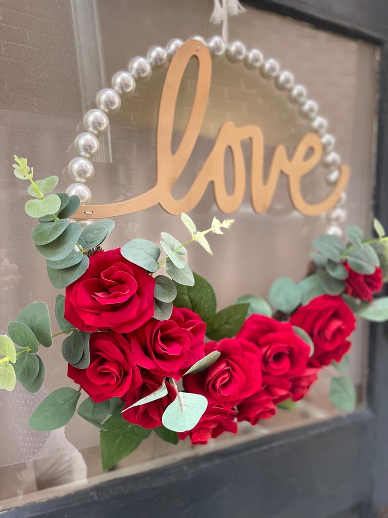 A close-up shot of a decorative wreath hanging on a glass door. The wreath features a circular base made of white pearls, with the word "love" written in a gold-toned script across the center. The bottom of the wreath is adorned with a lush arrangement of vibrant red roses and green eucalyptus leaves.