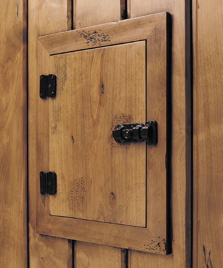 A close-up shot of a small wooden door built into a wood-paneled wall. The door features black metal hinges on the left and a matching black latch on the right, with a small decorative flourish etched into the top of the frame.