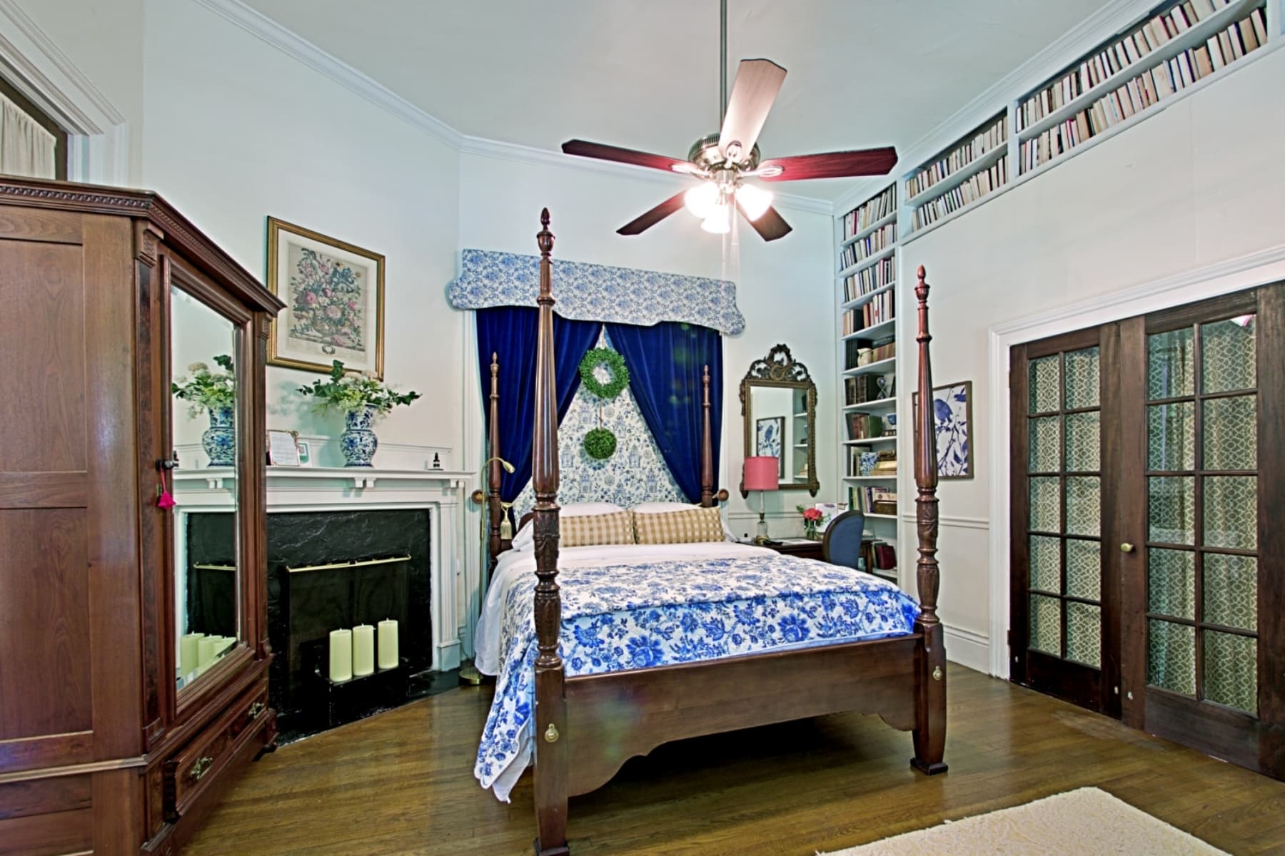 A high-angle view of a traditional bedroom featuring a dark wood four-poster bed with a blue and white patterned quilt. To the left, a large mirrored armoire stands near a white fireplace mantel decorated with greenery, while the right side of the room showcases a tall built-in bookshelf filled with books and a set of dark wood French doors.